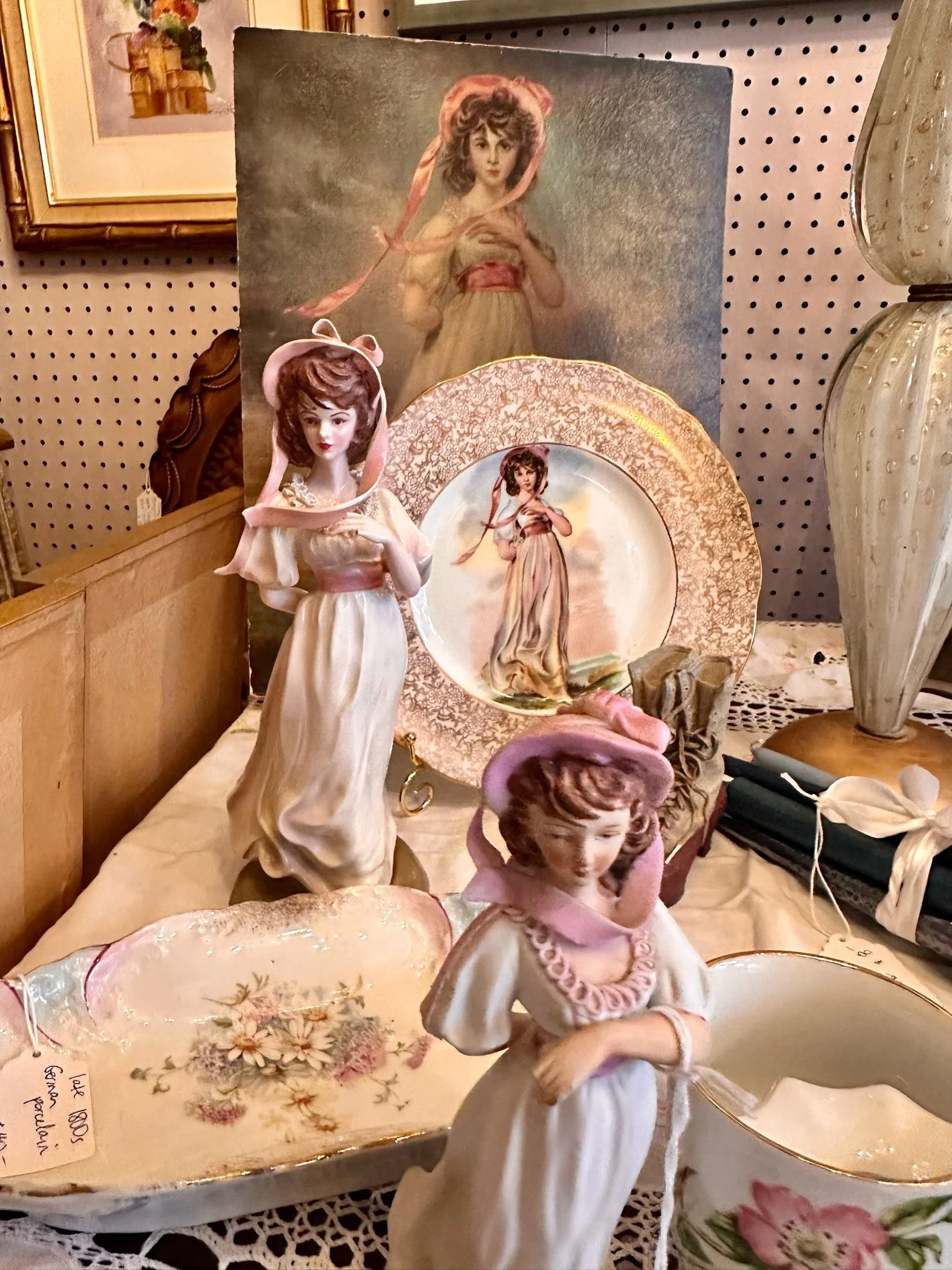 Inside Villa Vintiques, a display featuring Pinkie Girl. Collection of vintage porcelain figurines of women, decorative plates, and framed artwork, displayed on a table.