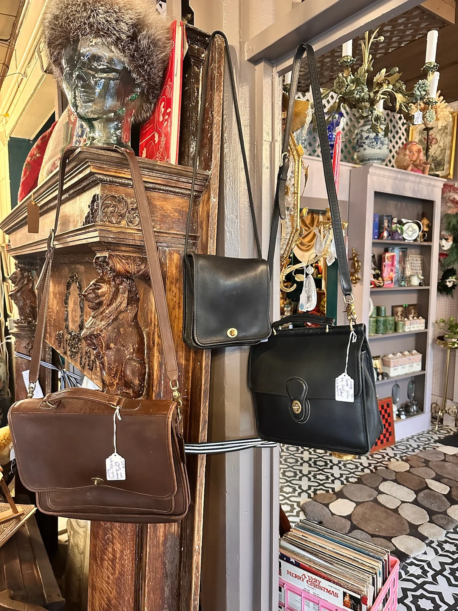Inside Villa Vintiques, a display of vintage handbags hung on a wooden antique fireplace mantle, with various decorative items and a bookshelf with records in the background.