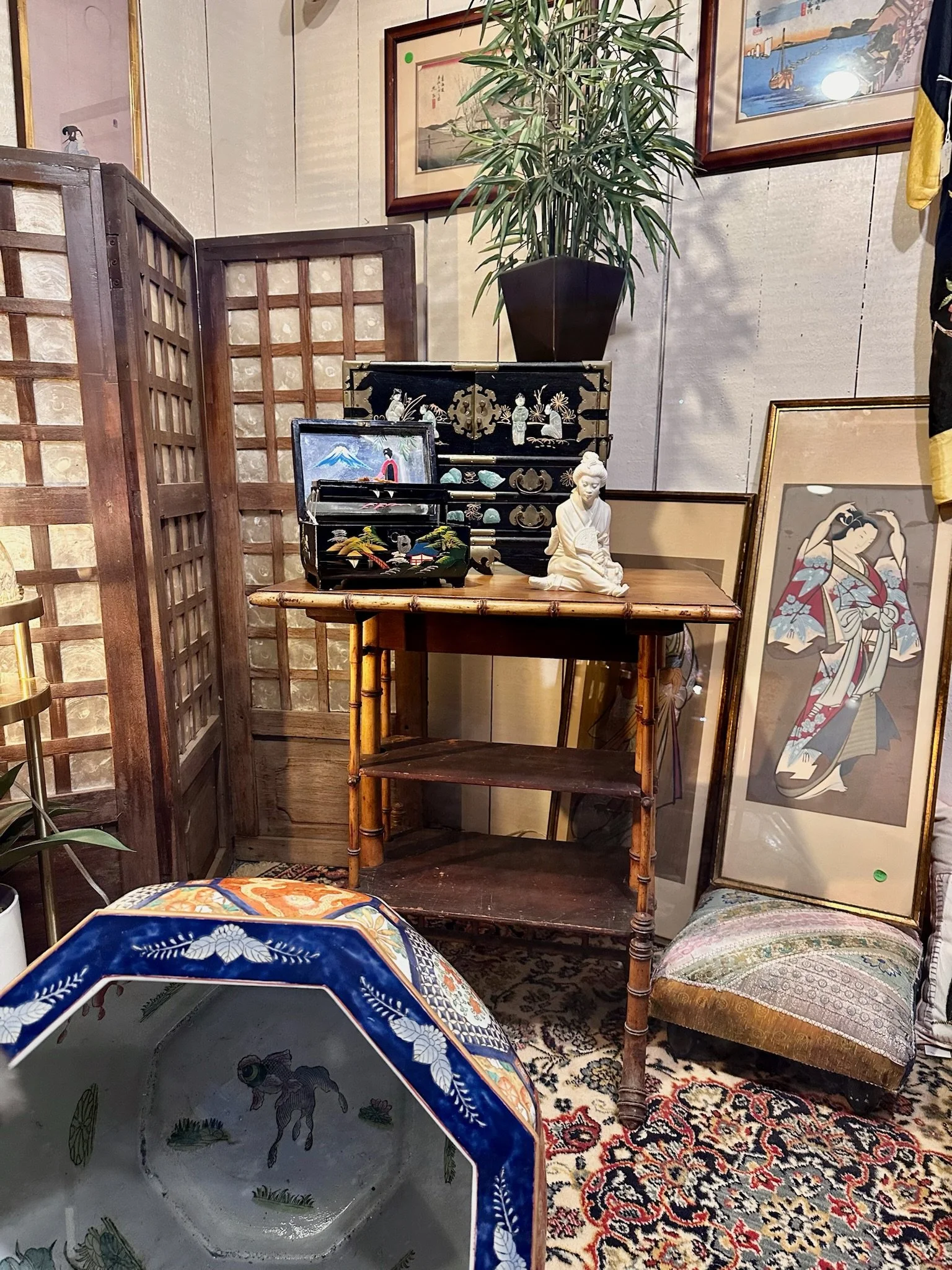 Villa Vintiques display window- a collection of Asian art and decor includes framed paintings on a wall, a large potted plant, and various decorative objects displayed on a wooden table and nearby furniture.