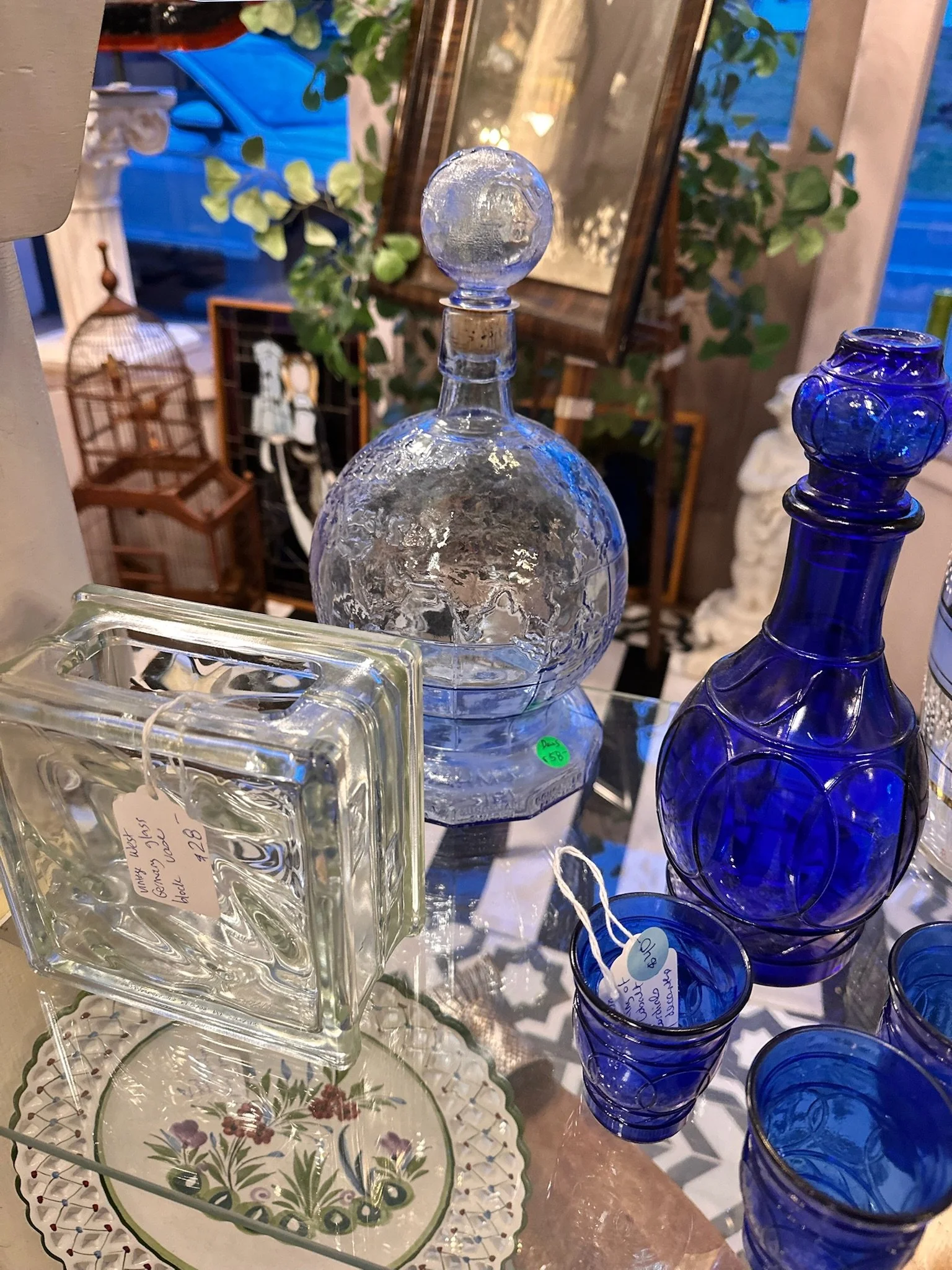Inside the tiled room at Villa Vintiques, a display of glassware including a large textured clear glass decanter with a ball-shaped stopper, and a stack of blue glass cups with a circular pattern.