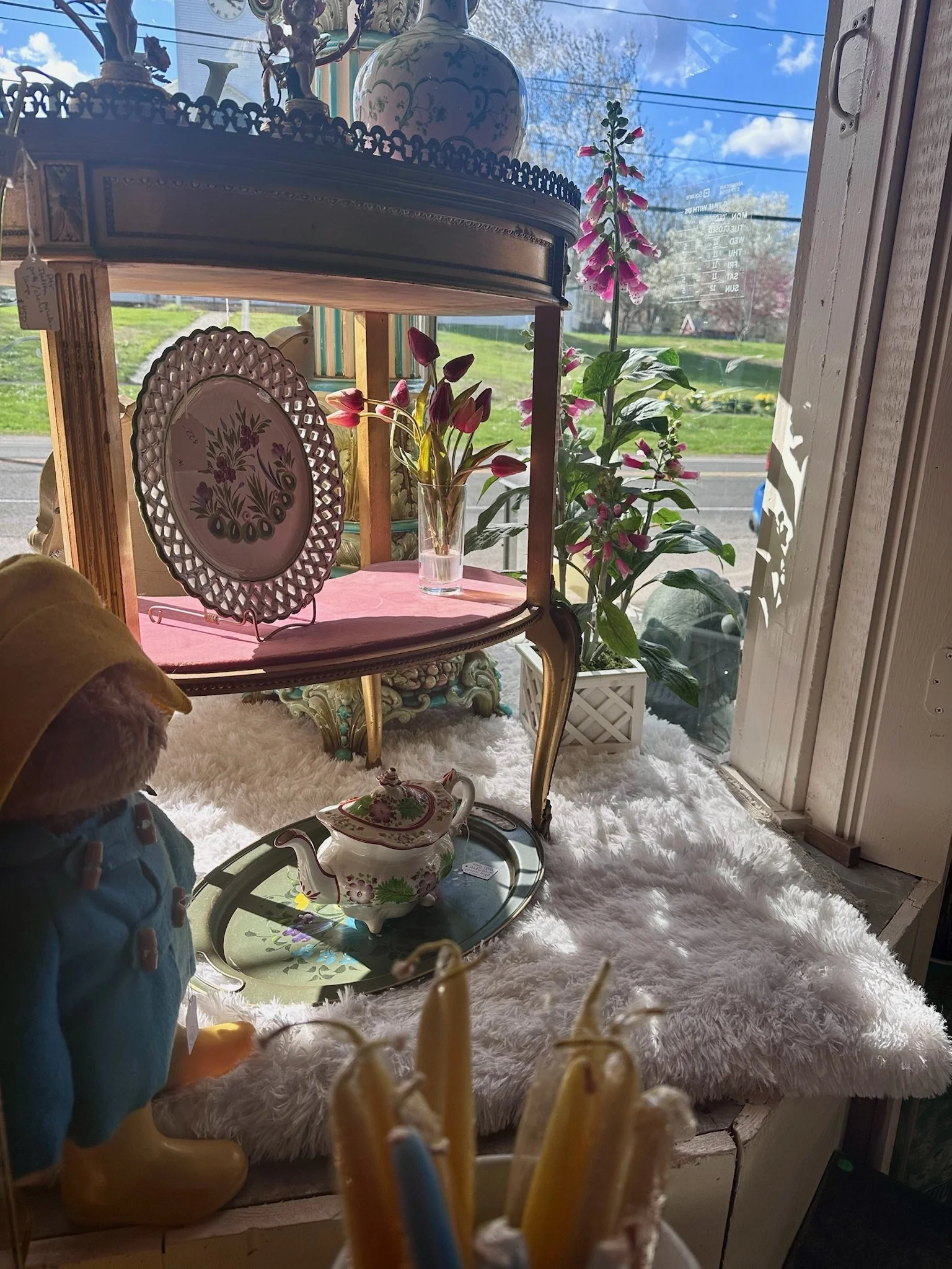 View from inside Villa Vintiques showing decorative objects including a porcelain vase on a glass and wooden shelf, pink flowers in a glass vase, a teapot, and a porcelain plate, with a window looking out onto a sunny day with blue sky and trees.