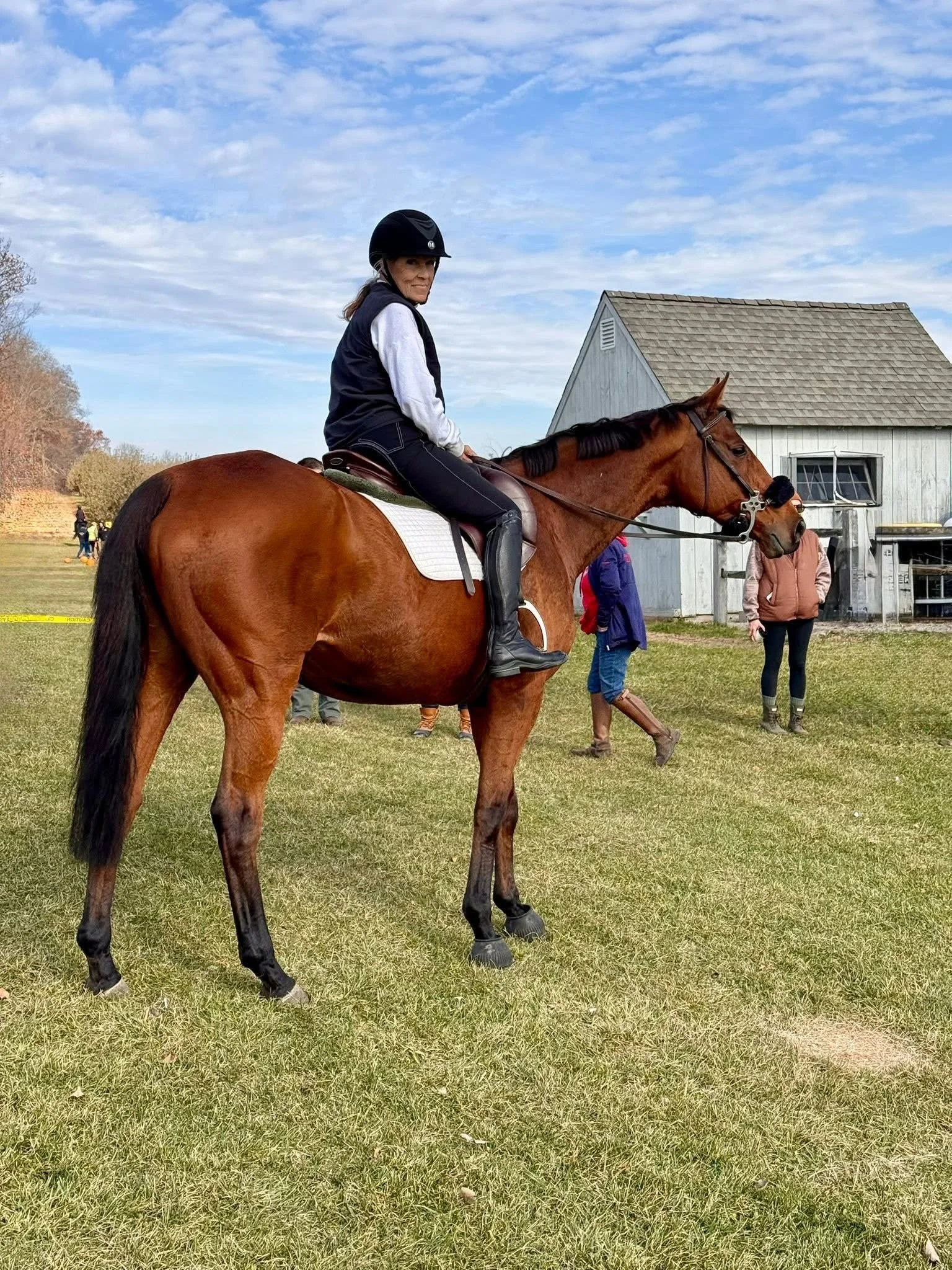 A woman riding a brown horse on a grassy field, wearing a riding helmet and a jacket, with a white barn and several people in the background.