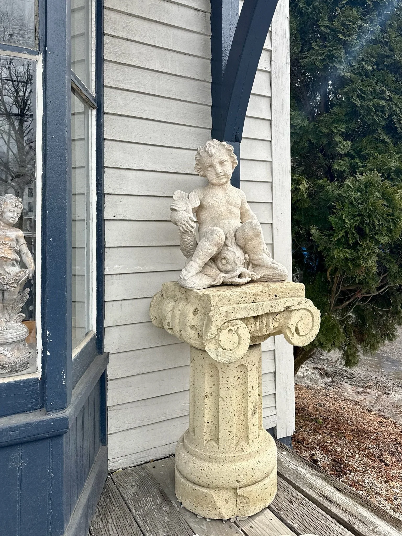 A stone sculpture of a cherub sitting on a turtle, mounted on a carved stone pedestal outside on the platform of Villa Vintiques with wooden siding and a window.