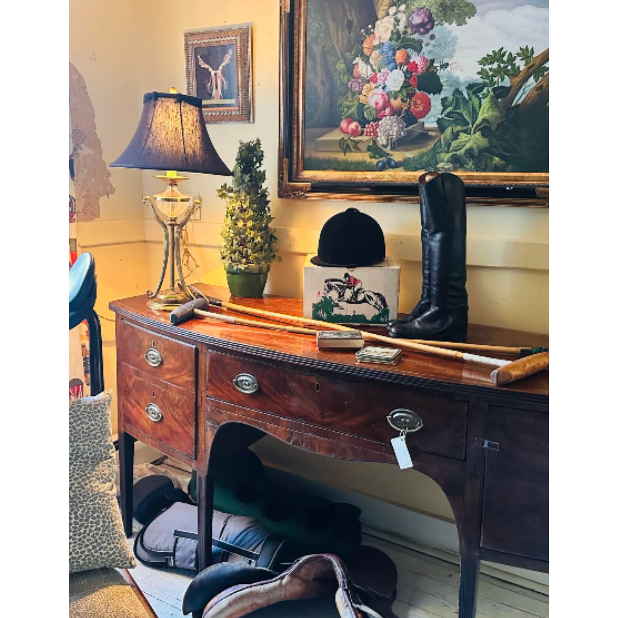 A wooden dresser in a room with multiple objects on its surface, including a brass lamp with a beige lampshade, a small potted plant, and a box with a zebra-patterned lid. A large, framed still life painting of a vase filled with colorful flowers han