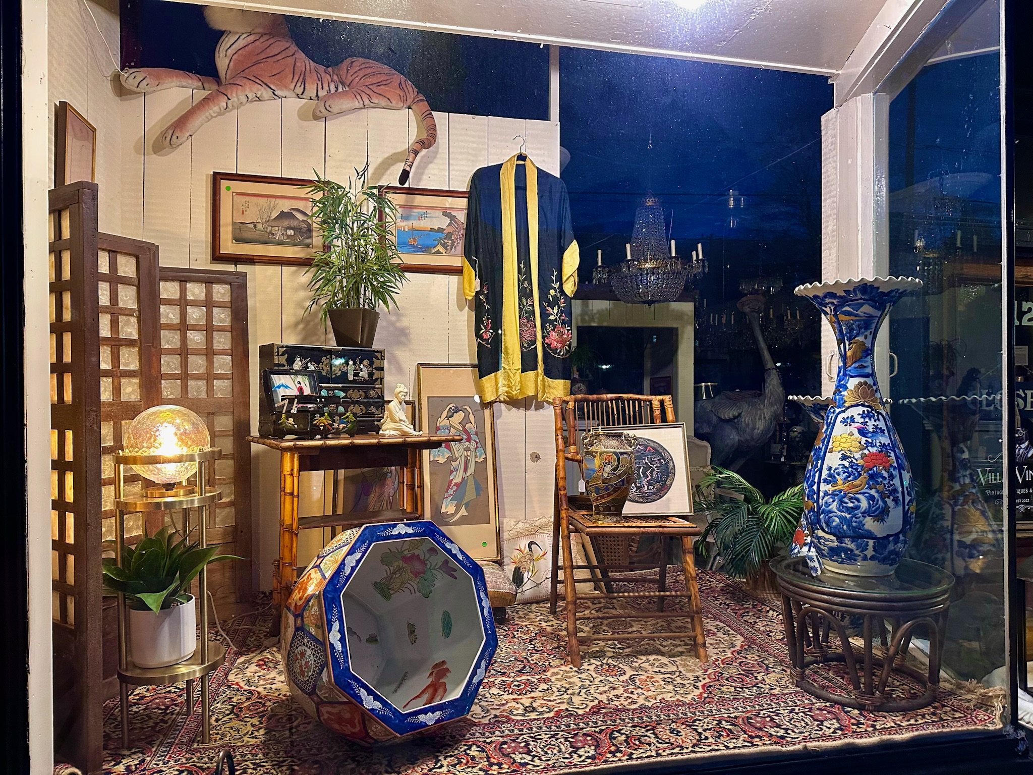 Villa Vintiques display window, filled with Asian art and antiques, including framed paintings, a porcelain vase, wooden chairs, a gilded table, a leopard print stuffed animal, a tall plant, a chandelier, and brightly patterned textiles.
