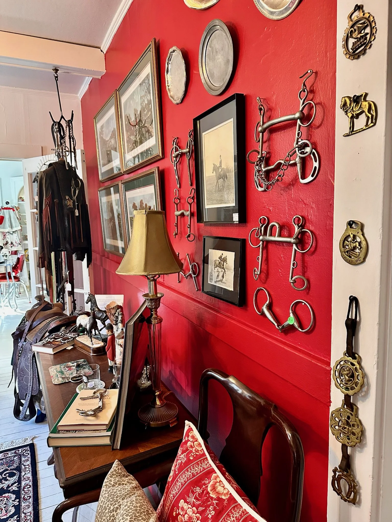 Inside The Silver Spur Equestrian Consignment room at Villa Vintiques. A red wall decorated with horse bits, framed pictures, and a picture of a person riding a horse. A wooden table below holds books, jewelry, and decorative items.