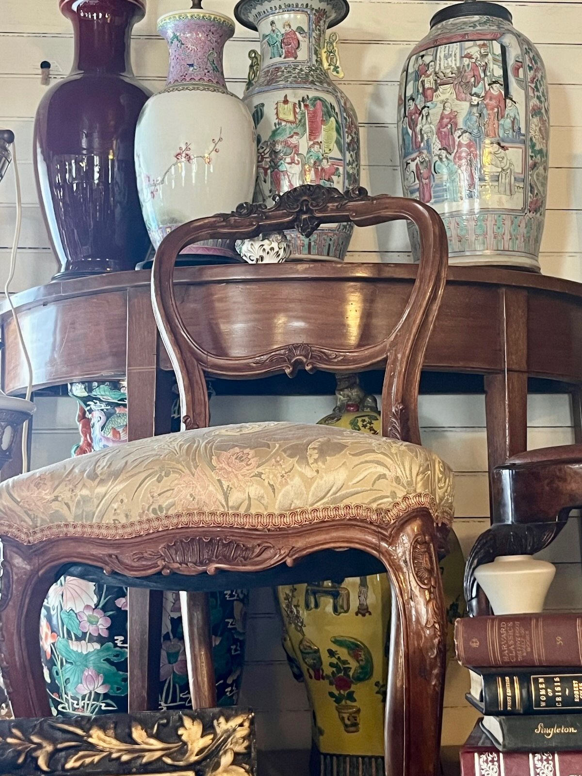 Inside Villa Vintiques, a collection of decorative vases and antique furniture, including a wooden chair with an ornate carved back and a cushioned seat, displayed against a wooden-paneled wall.