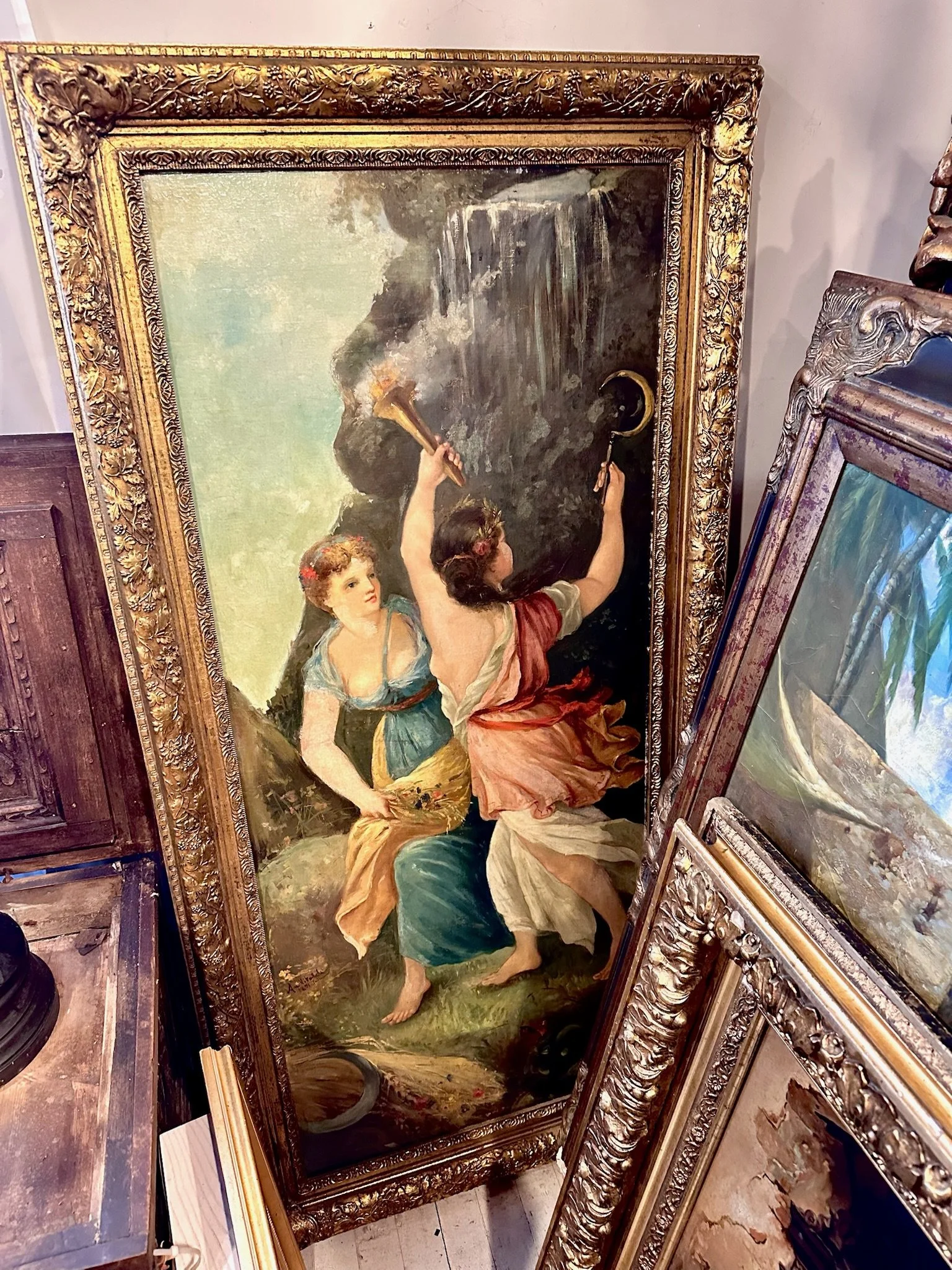 Some of the art for sale at Villa Vintiques. A painting of two women in a natural outdoor setting, one holding a horn and the other with an intricate hairstyle and gown, surrounded by ornate gold frames.