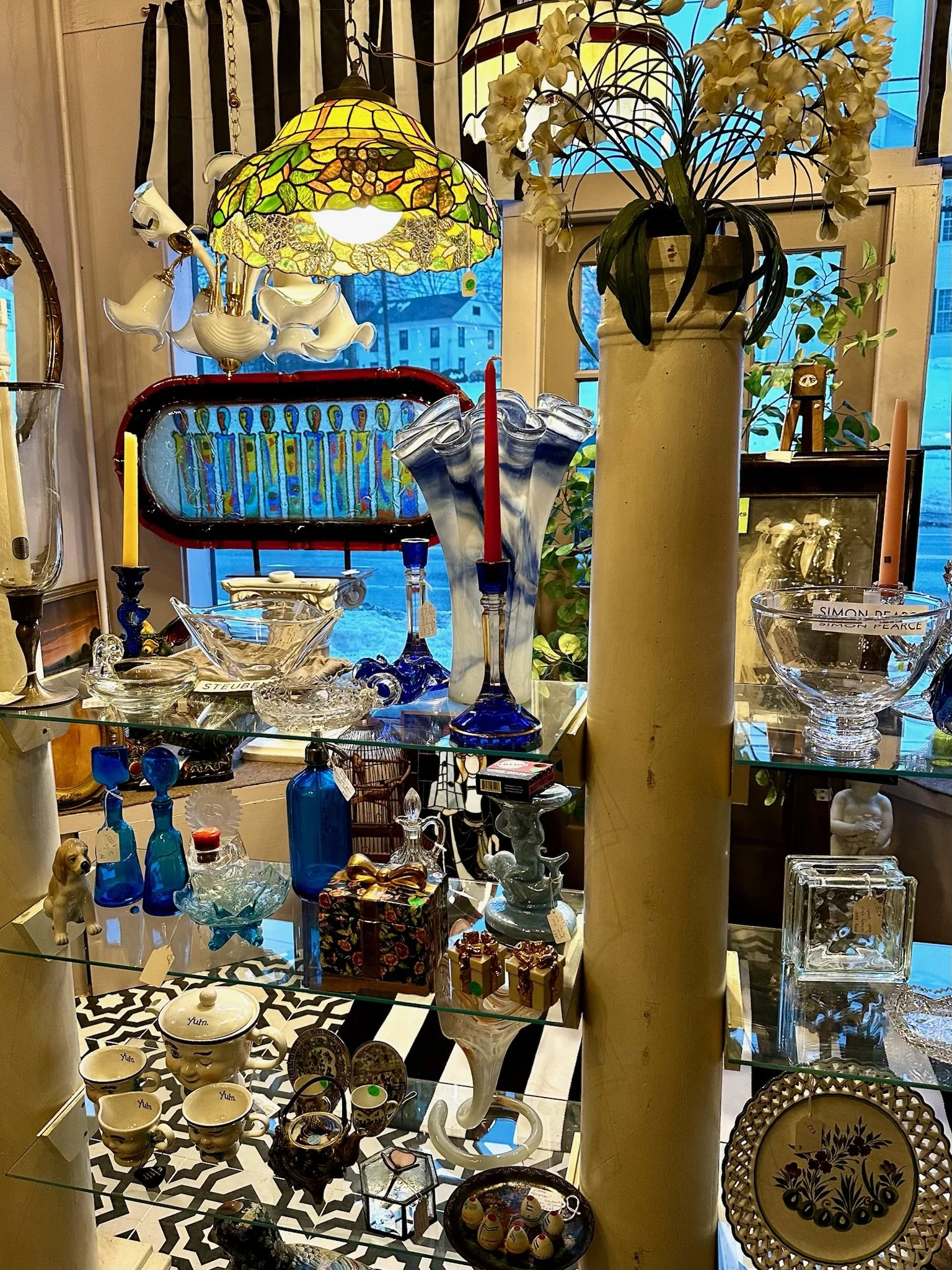 Inside Villa Vintiques, a display of decorative glassware, vases, and ceramics on glass shelves, with a stained glass lamp, a vase with white orchids, and framed pictures in the background.