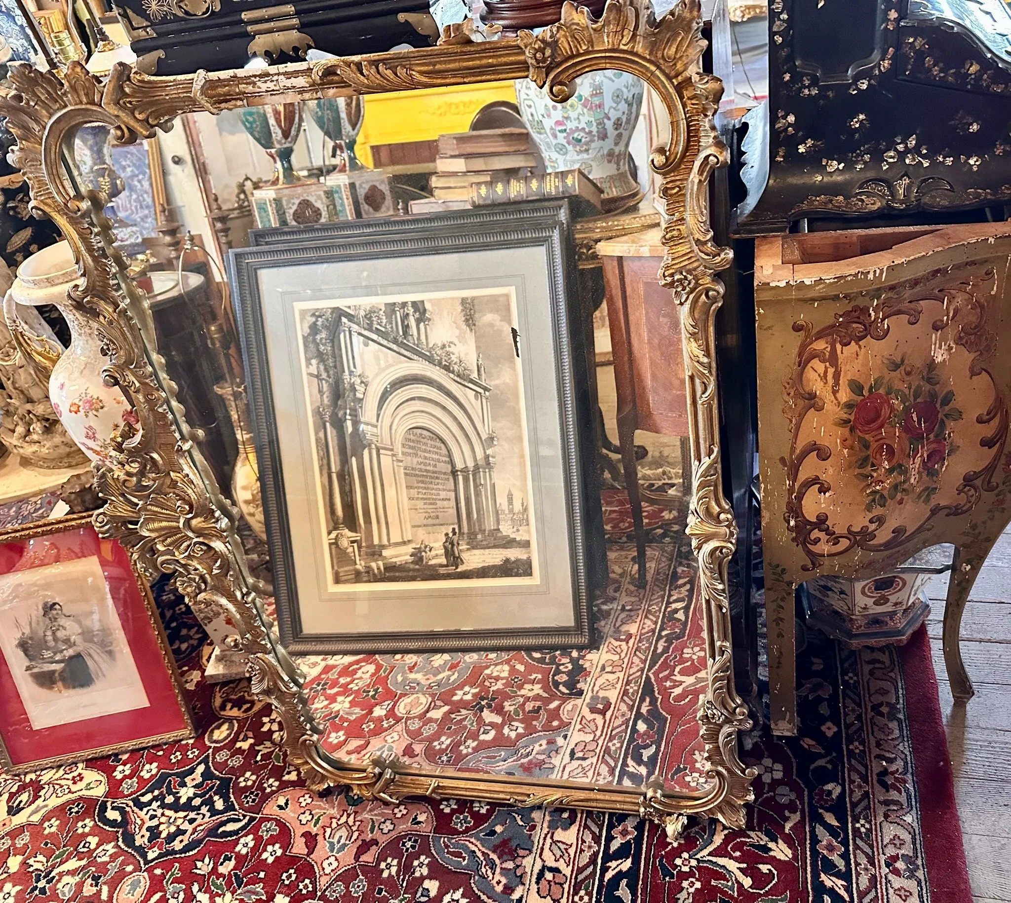 Inside Villa Vintiques, an ornate gold-framed mirror leaning against a gilded decorative stand, with framed artwork and various decorative items on a table and shelves in the background.