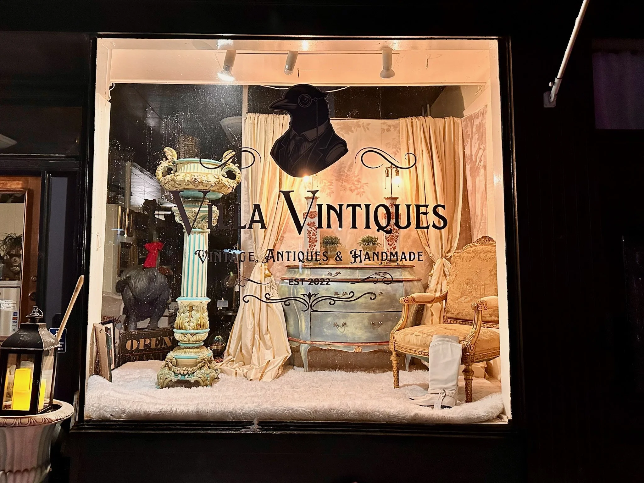 Storefront window display with vintage furniture, a bust, and decorative items inside, sign reading 'Vila Vintiques Vintage, Antique & Handmade'.