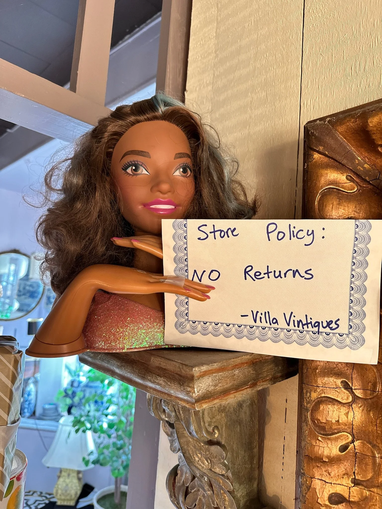 A Barbie head doll with curly brown hair, brown skin, and painted makeup, holding a sign that reads "Store Policy: NO Returns" at Villa Vintiques.