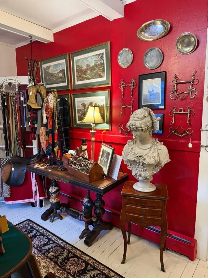 Interior of an antique shop with a red wall decorated with framed landscape paintings, silver trays, and vintage hardware. A bust sculpture of a woman sits on a wooden side table, and a vintage table lamp provides light. A dark wooden table displays 