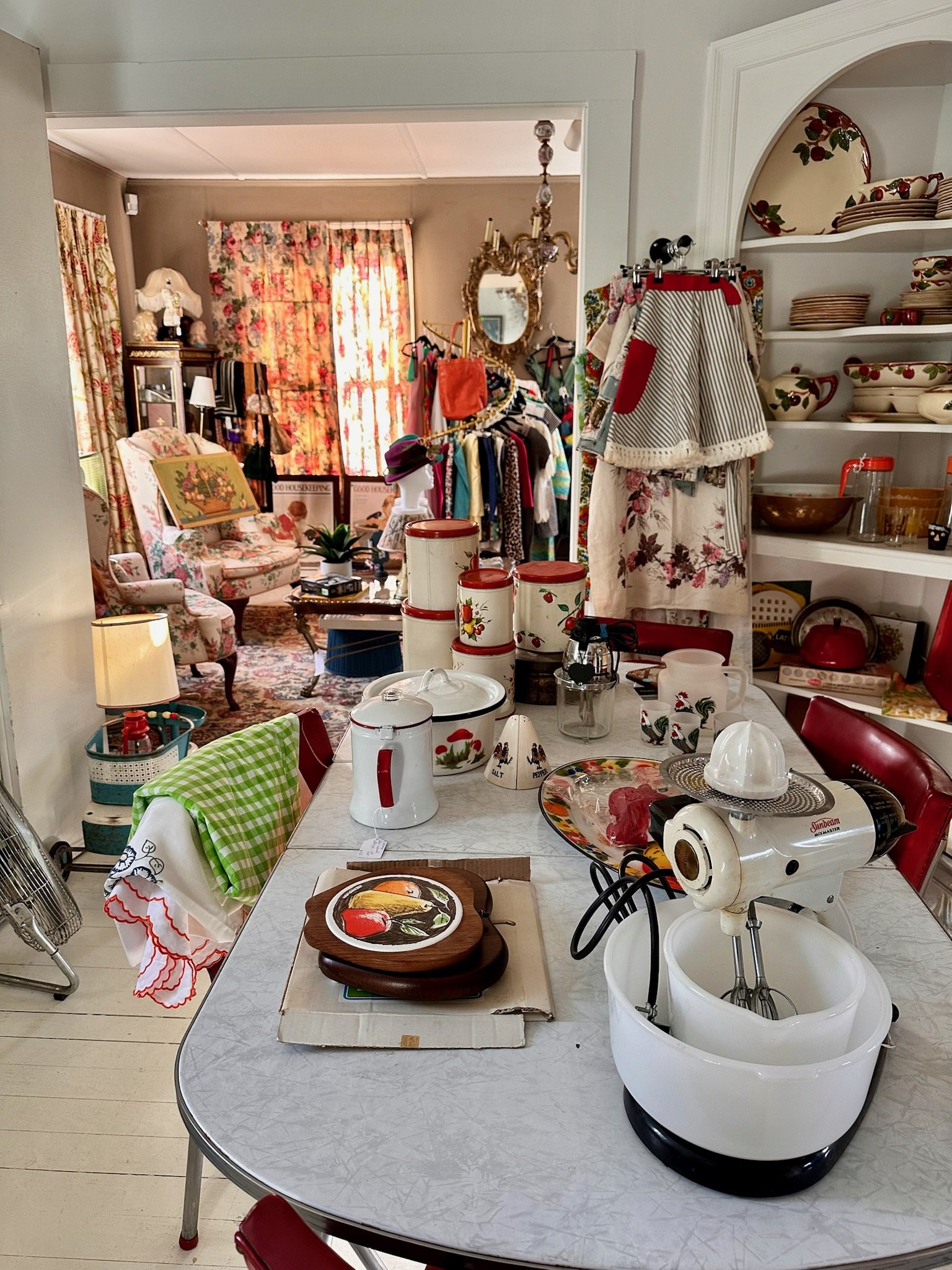 The mid-century room upstairs at VV, filled with vintage dishware, textiles, and kitchen appliances. In the foreground, a white table holds a white stand mixer, a white collecting jar, a decorative roaster, and a tray with a painting of fruits.