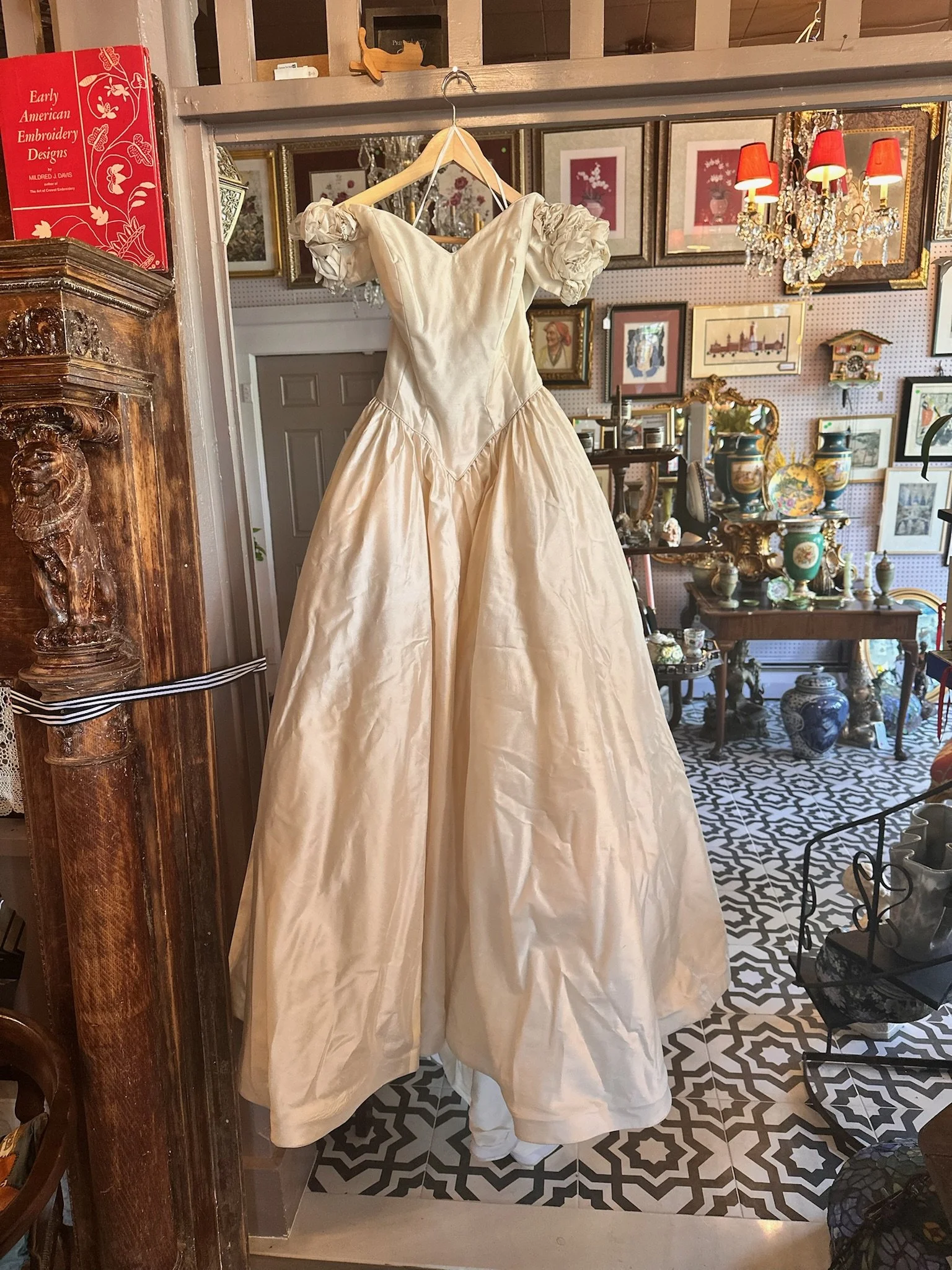 A vintage wedding gown with puffed sleeves hanging on a wooden hanger inside Villa Vintiques with framed artwork, a chandelier, and decorative vases in the background.