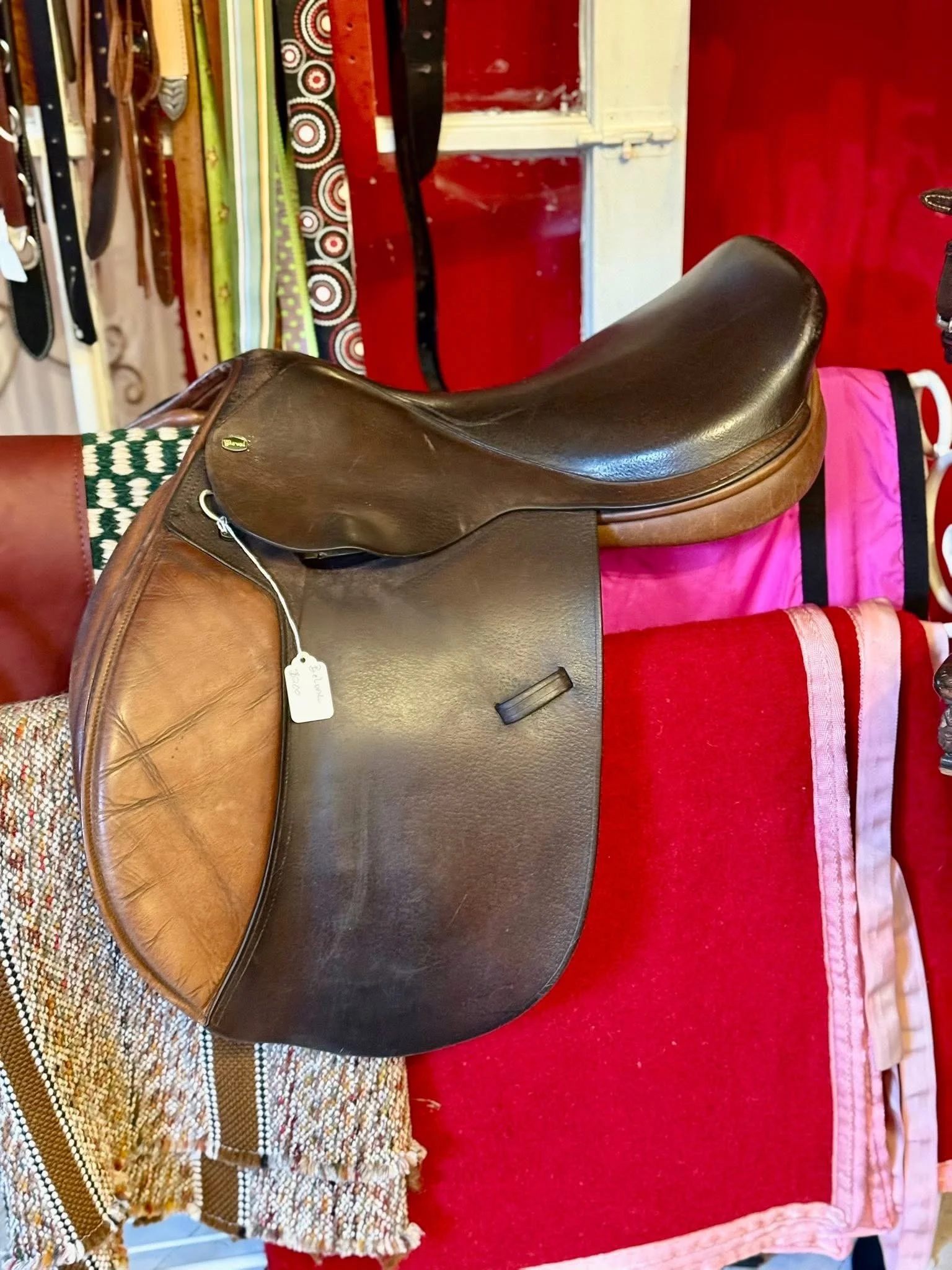 A brown leather saddle on a red tablecloth with a multicolored woven fabric underneath. In the background, there's a shelf with belts and straps, and various fabrics with patterns and colors.