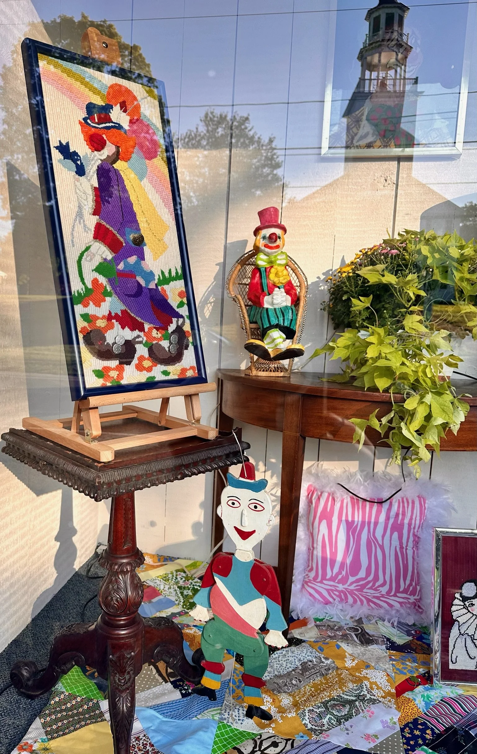 Villa Vintiques Store Window Display of colorful decorative art pieces including a painted clown doll, and a painted paper clown figure, arranged on a wooden table with a window showing reflections of a house and trees outside.
