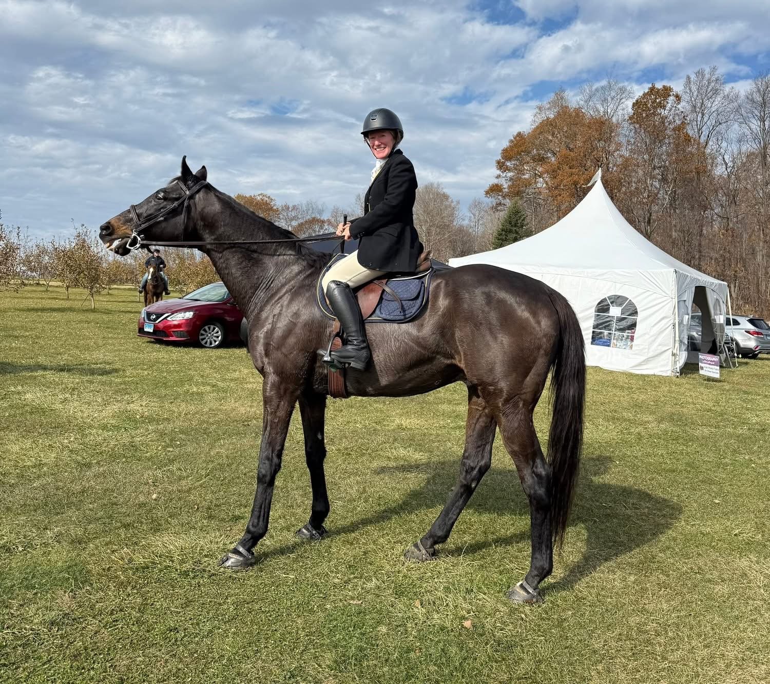 A woman in equestrian riding attire, including a black helmet, jacket, and riding boots, riding a dark brown horse on a grassy field. In the background, there is a white event tent, a red car, and some trees with autumn foliage.