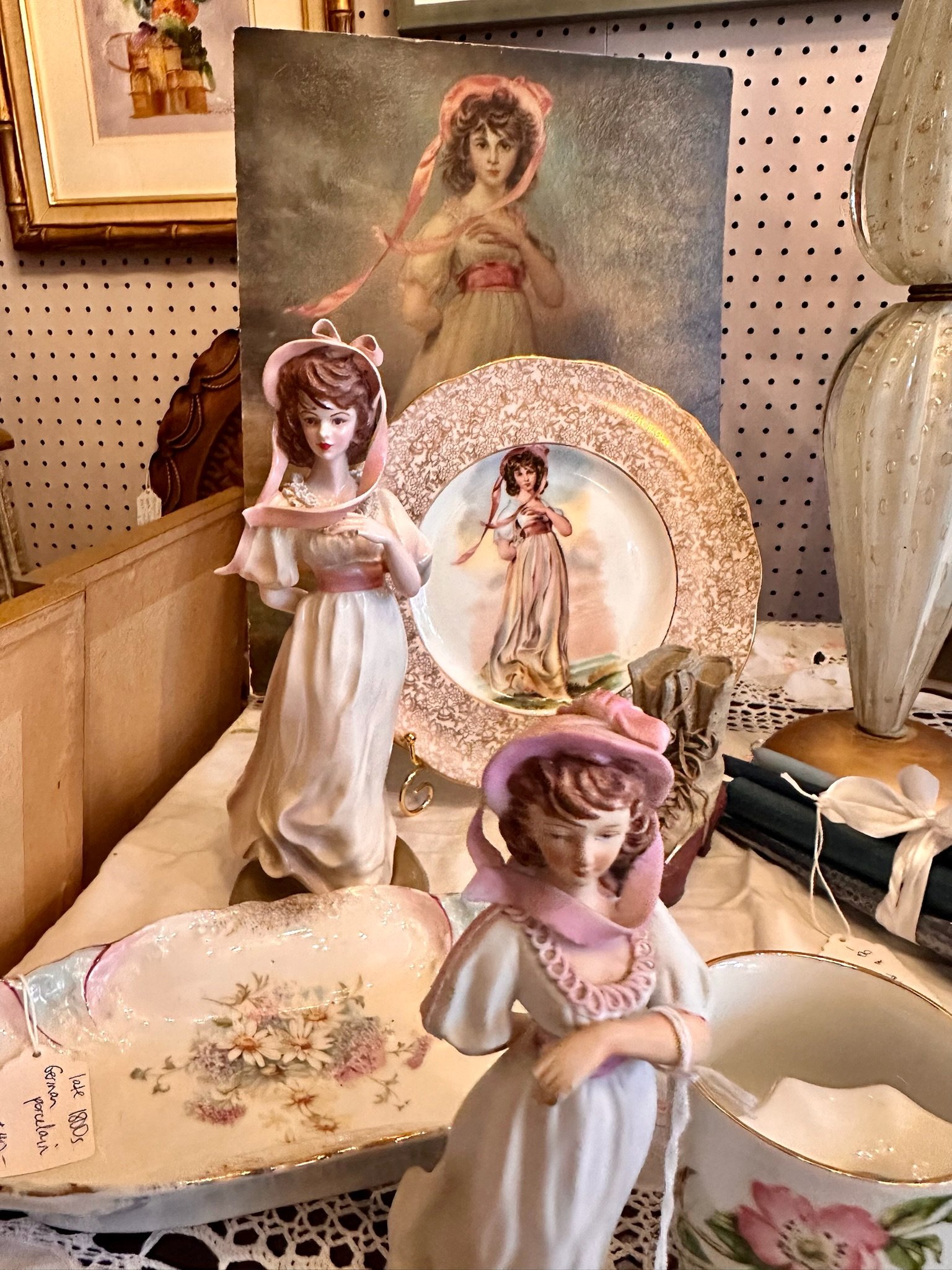 Collection of vintage porcelain figurines including young women and girls, decorative plates, and pastel-colored ceramics displayed on a table.