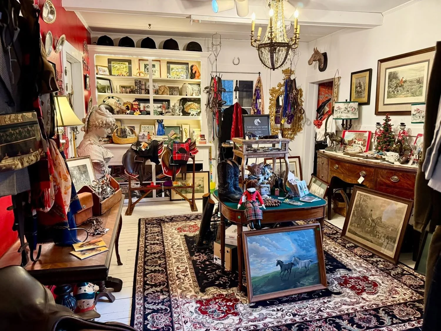 A cluttered antique shop with various framed pictures, artworks, and decorative items displayed on shelves and furniture. The room features a Persian rug, vintage lamps, and a chandelier. Items include horse paintings, a mannequin bust, dolls, hats, 