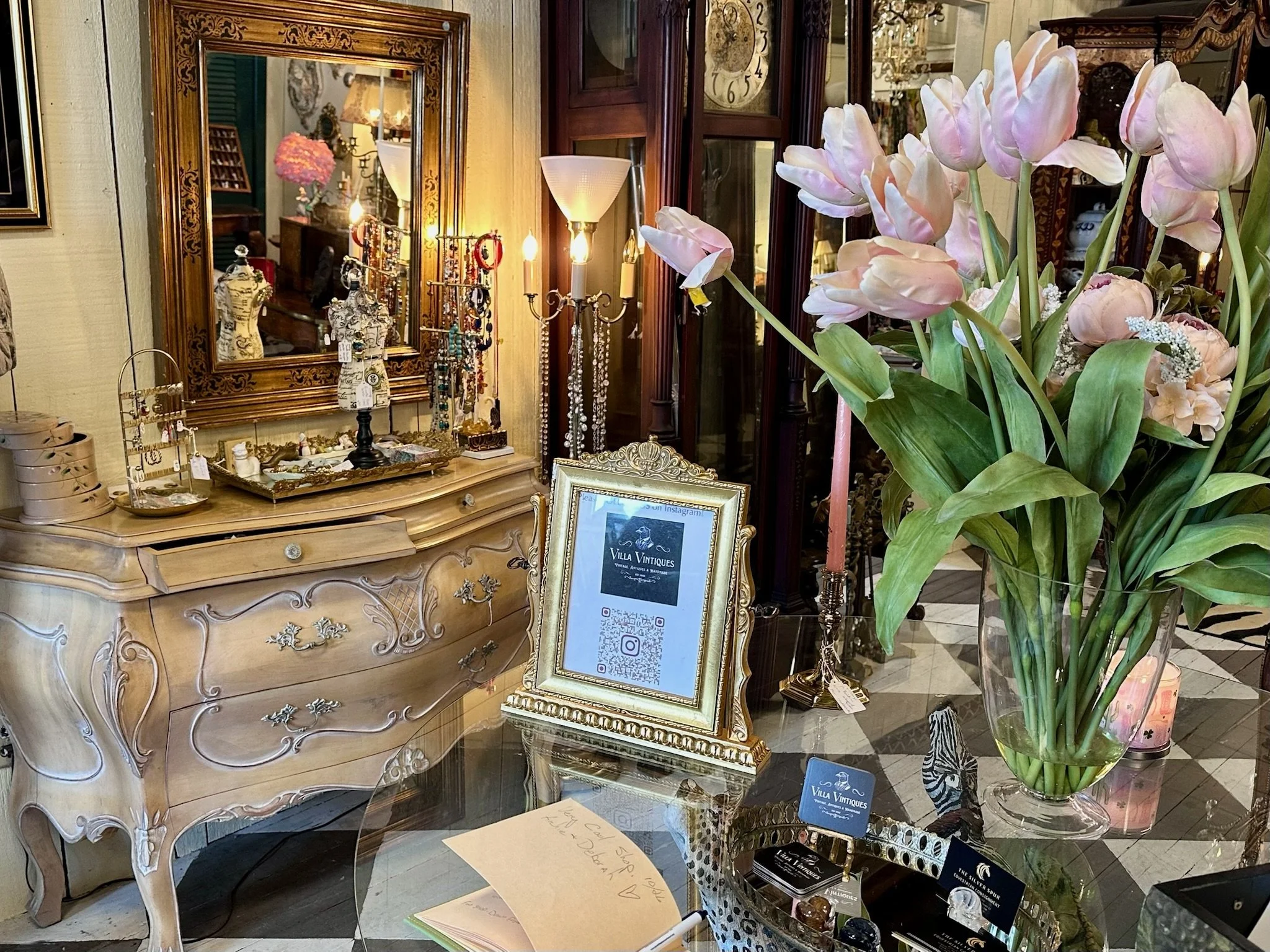 Decorative jewelry display with necklaces and earrings on a stand, ornate mirror above a vintage dresser, large bouquet of pink and white tulips in a tall glass vase, framed sign with QR code, and other decorative items inside Villa Vintiques.