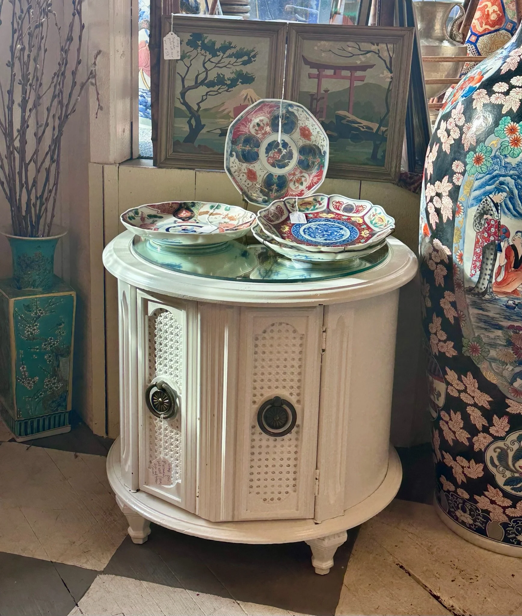 Display of decorative Japanese-style plates and paintings, including a landscape with a tree and a torii gate, on an antique white furniture piece in Villa Vintiques.