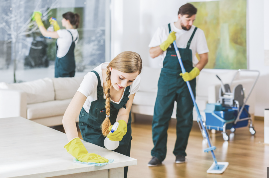 Three people cleaning a living room: a woman wiping a table, a man mopping the floor, and another woman cleaning a window, all wearing gloves.