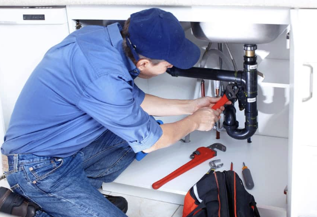 A plumber working under a kitchen sink, using a pipe wrench to tighten or loosen pipes.