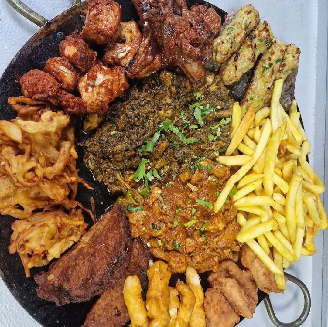 Assorted Nigerian dishes including fried chicken, goat meat, plantains, French fries, and a vegetable stew served on a black platter.