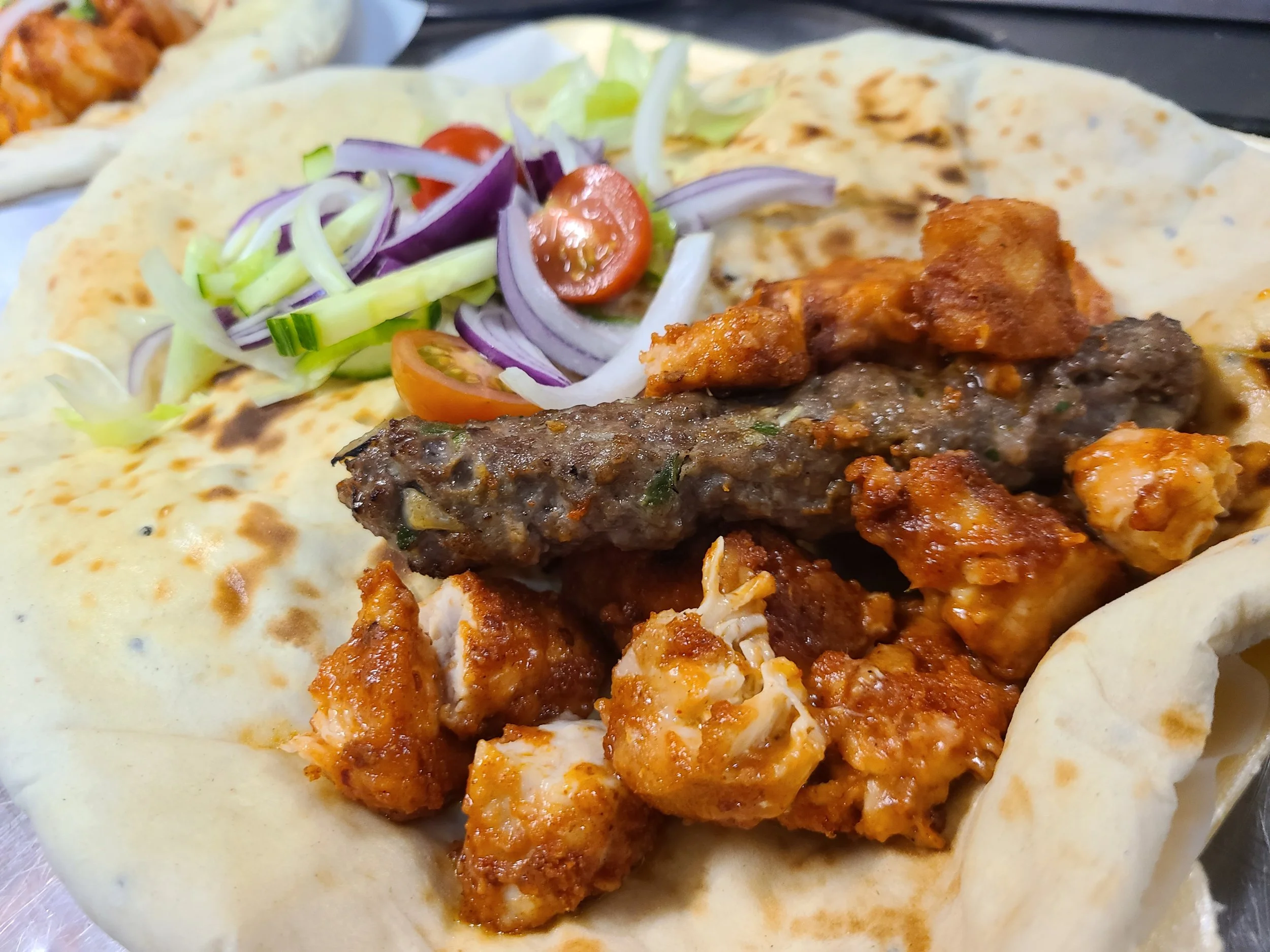 Close-up of a breakfast taco with grilled flatbread, topped with shredded lettuce, sliced cherry tomatoes, thinly sliced onions, and seasoned grilled beef and chicken pieces.