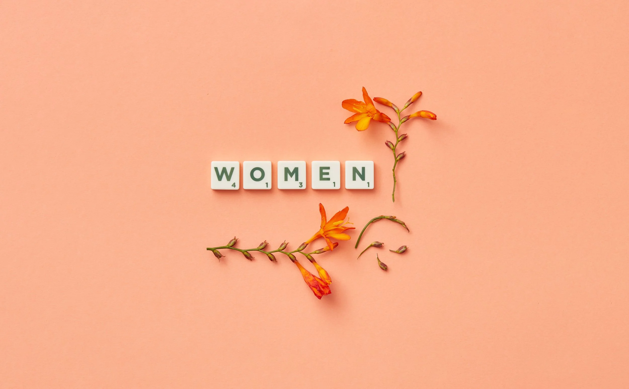 she breathes again pdx. menopause coaching Pink background with the word 'WOMEN' spelled out in white Scrabble tiles, and orange flowers and green vine arranged around the tiles.