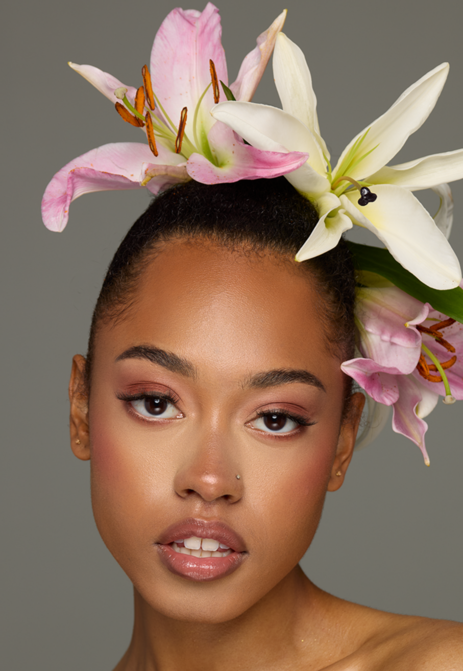 Portrait of a woman with a floral headpiece featuring pink and white lilies, wearing makeup, and having a neutral expression against a plain background.
