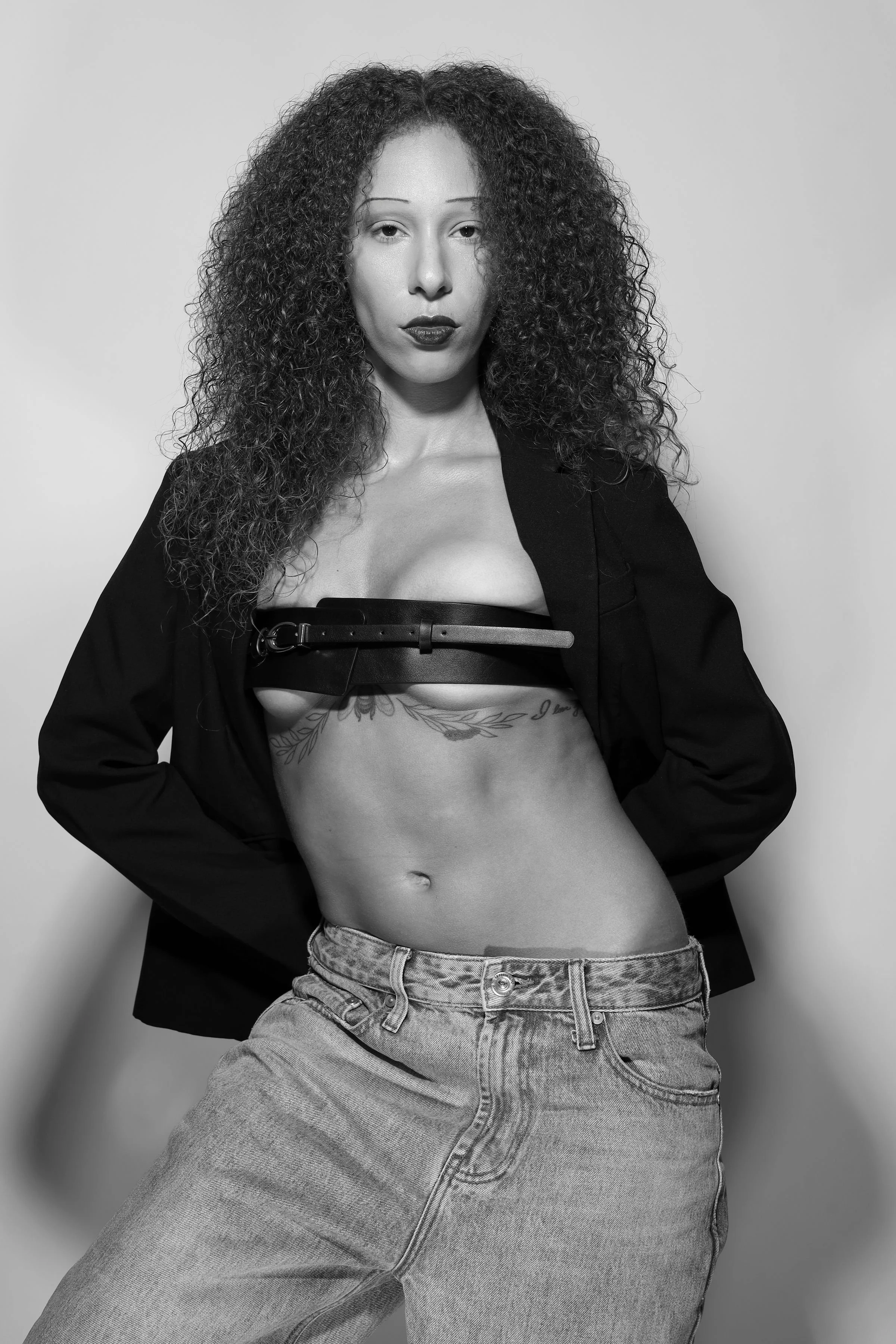 Black and white photo of a woman with curly hair, wearing a black blazer only partially covering her shoulders, a belt, and jeans, with some tattoos visible on her torso, standing against a light plain background.