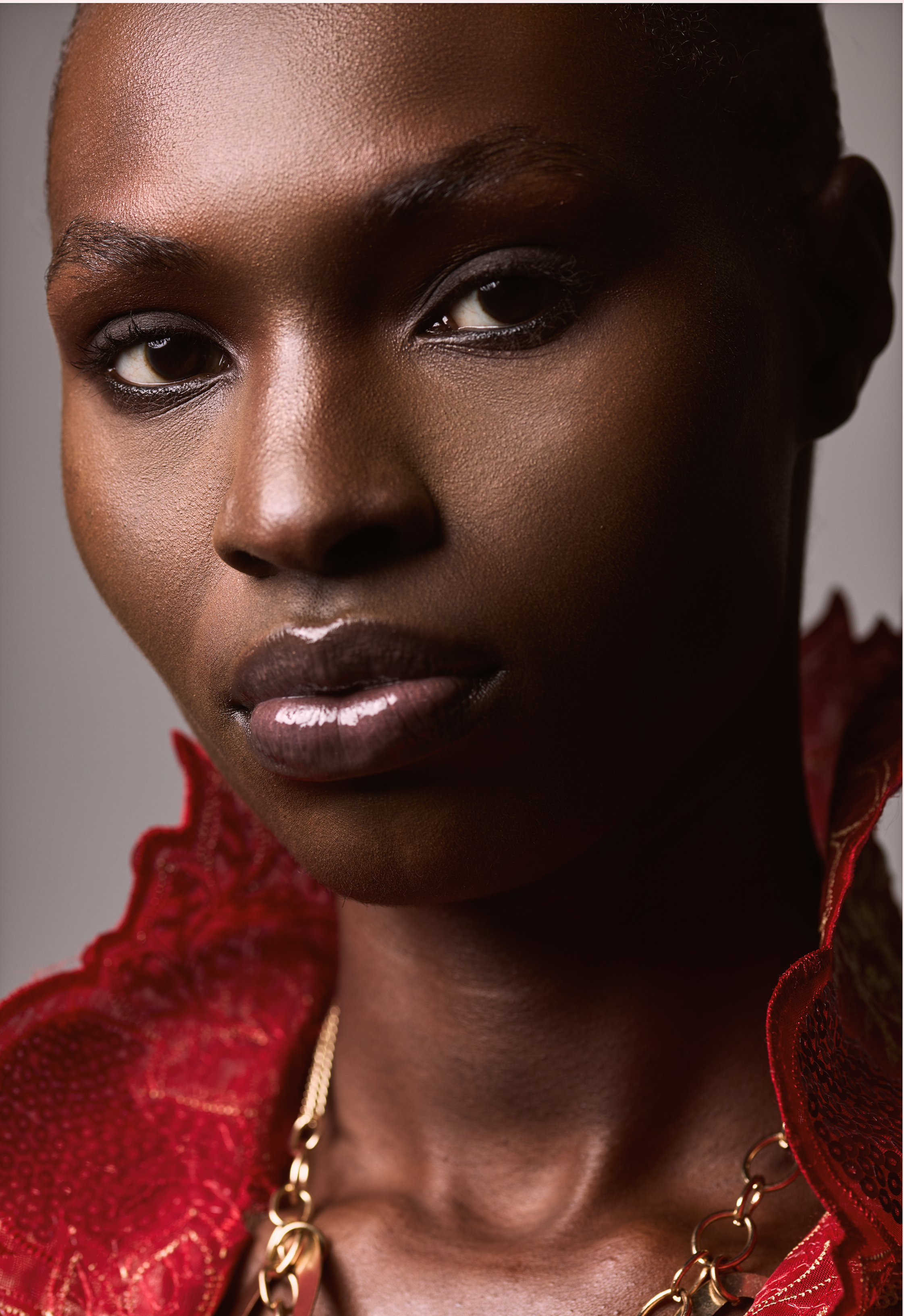 Close-up of a woman's face, showing her dark skin, eye makeup, full lips with glossy lipstick, wearing a red lace clothing with gold chain necklace.