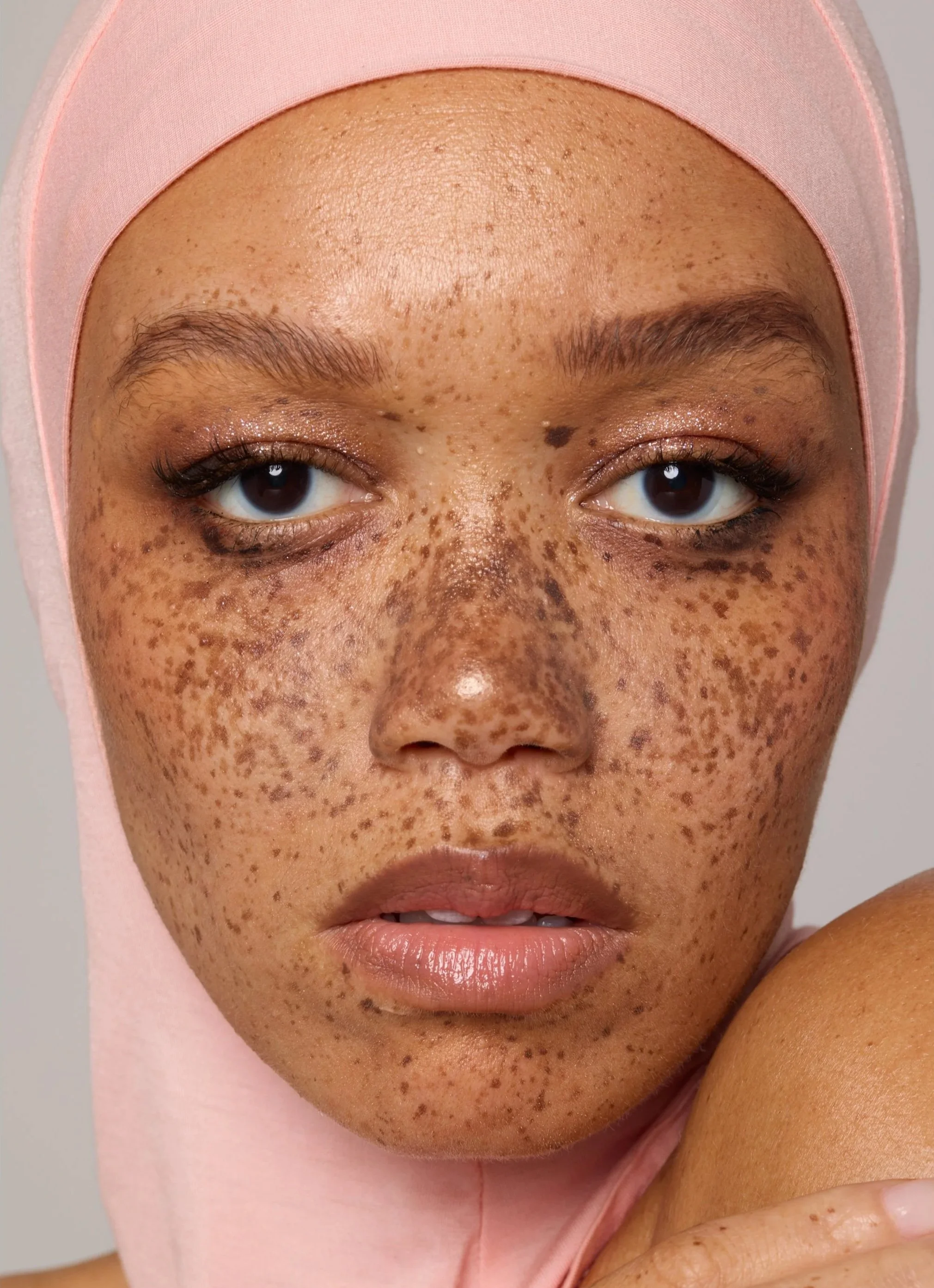 Close-up of a woman's face showing her brown skin with freckles and dark eyes, wearing a soft pink hijab.