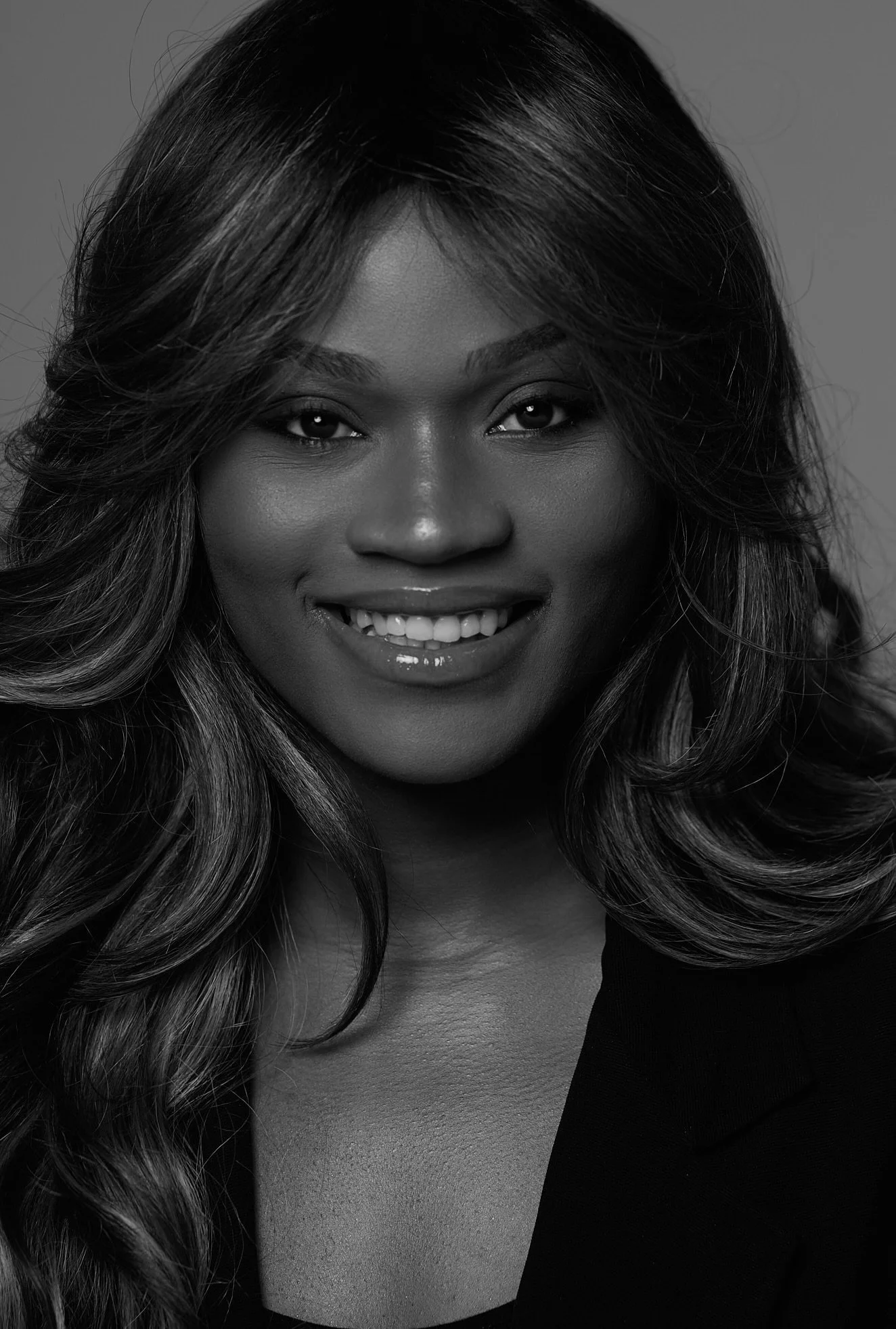 A black-and-white close-up portrait of a smiling woman with wavy hair, wearing a dark top with an off-the-shoulder style.