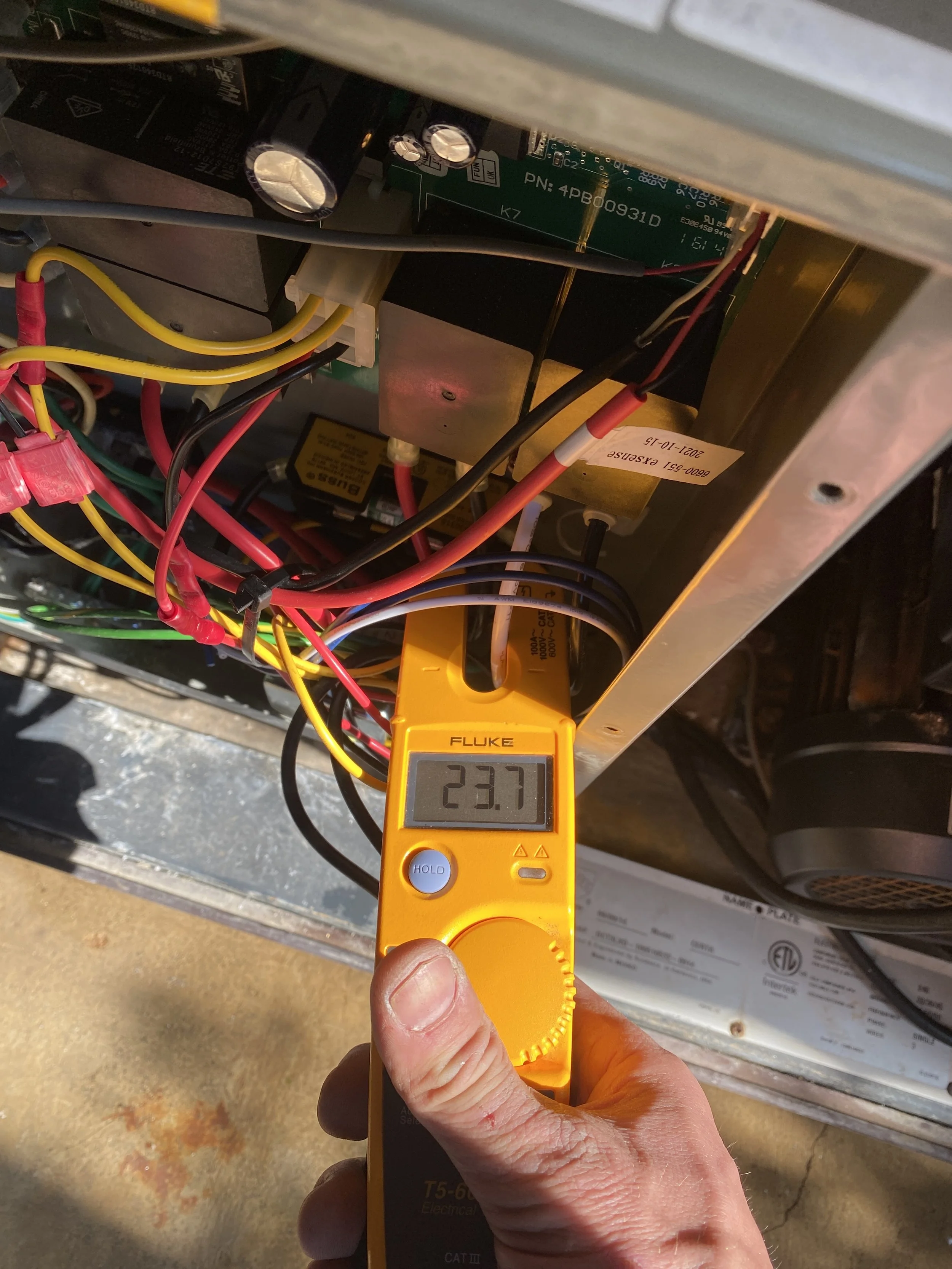 Person holding a digital multimeter with a reading of 23.7, testing inside an electronic device with multiple wires and circuit boards.