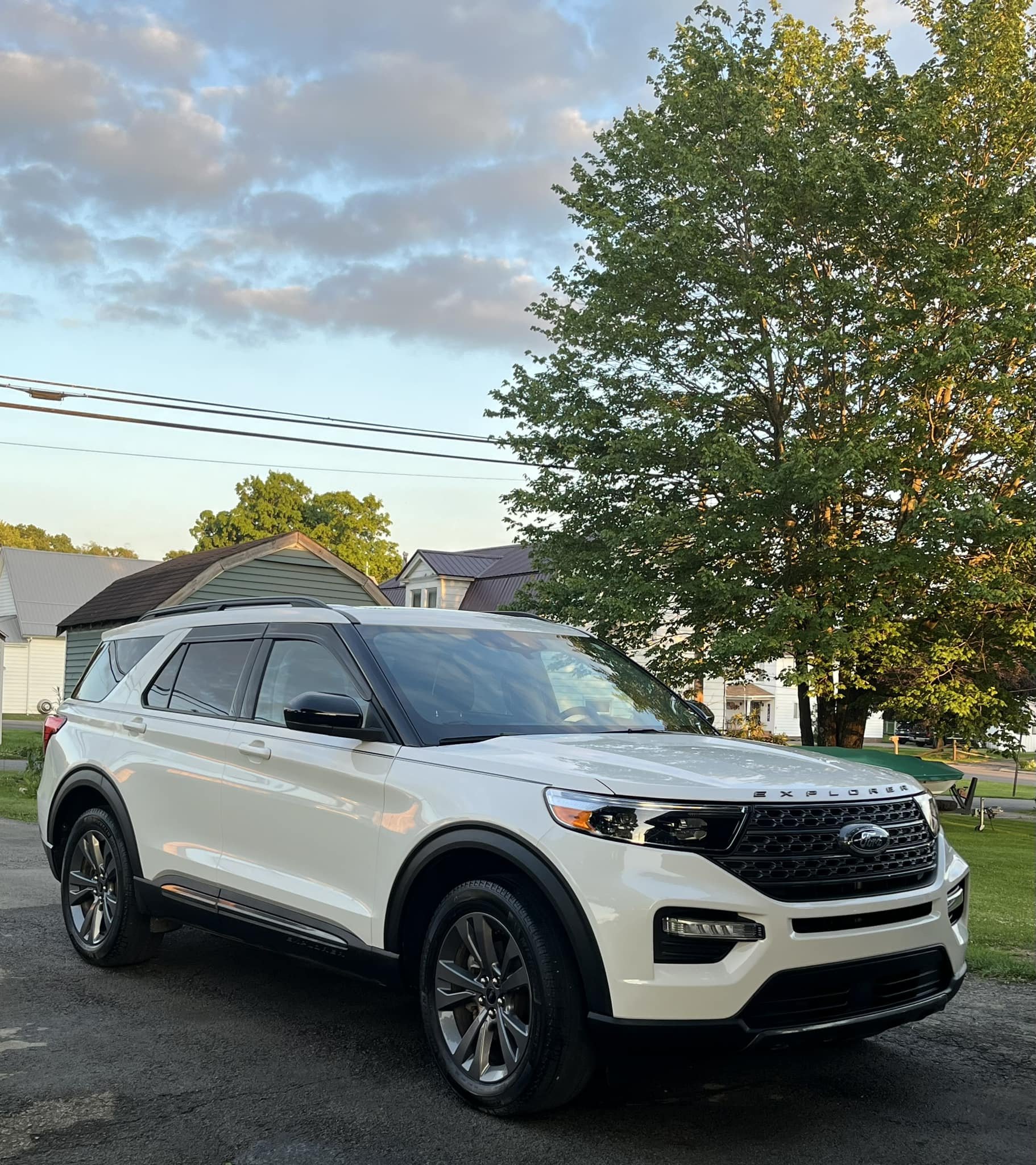 A white Ford Explorer SUV parked on a paved surface under a partly cloudy sky, with trees and houses in the background.