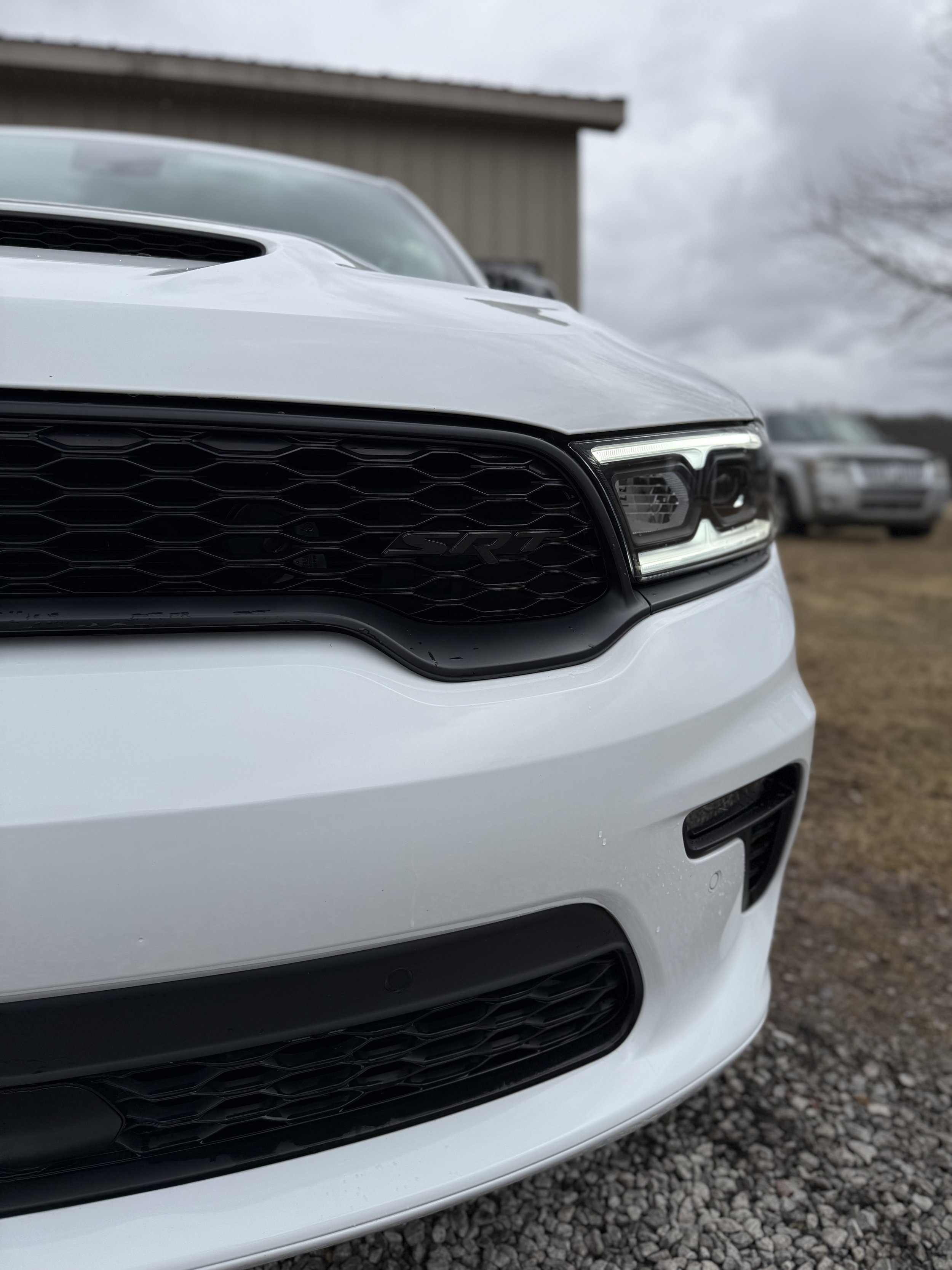 Close-up of the front part of a white sports car, showing the grille with a honeycomb pattern and the headlight, parked on gravel with a building and cloudy sky in the background.