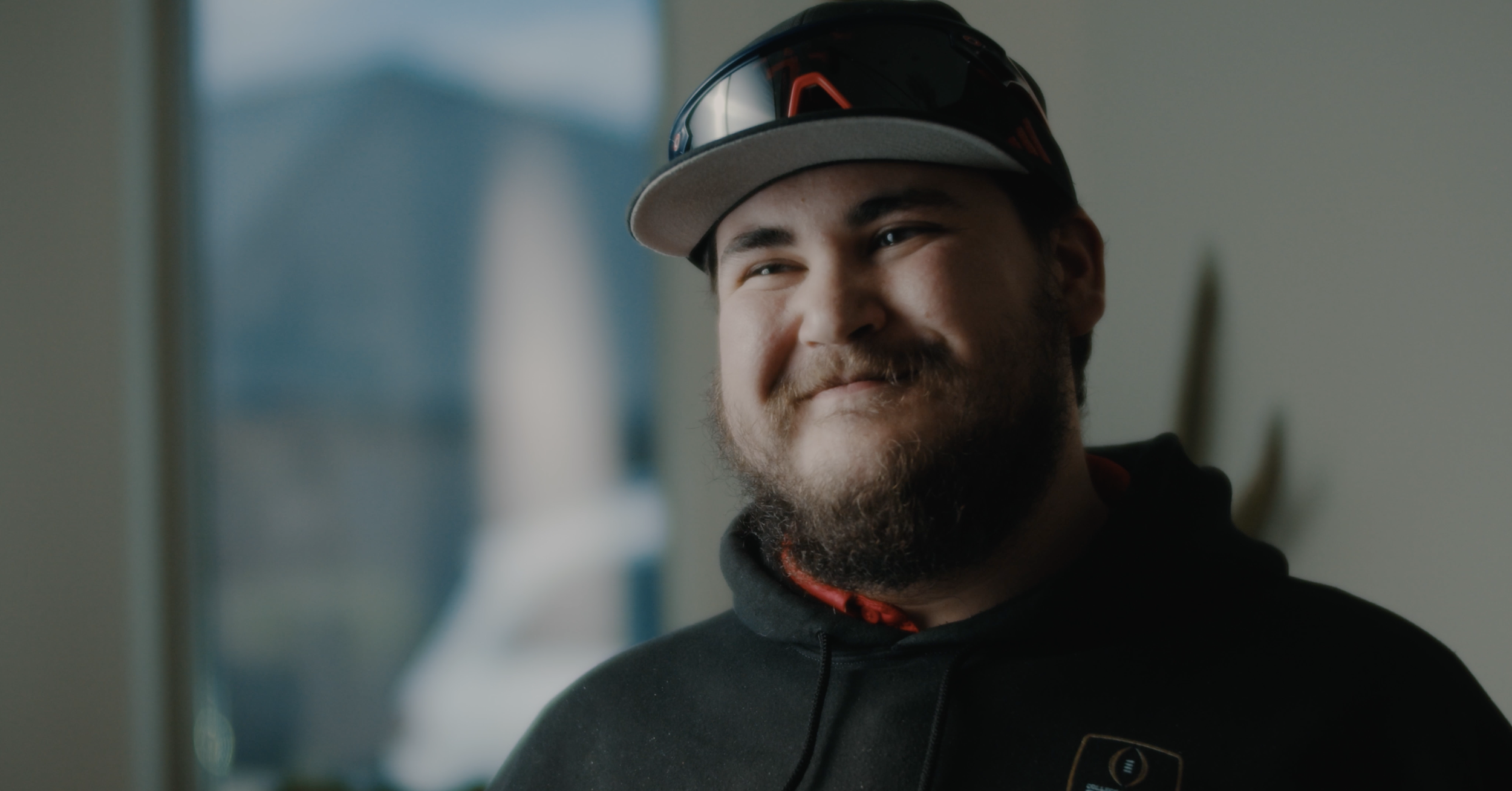 Patient Testimony | Close-up of a smiling man with a beard wearing a black hoodie, cap, and sunglasses on his head, indoors.