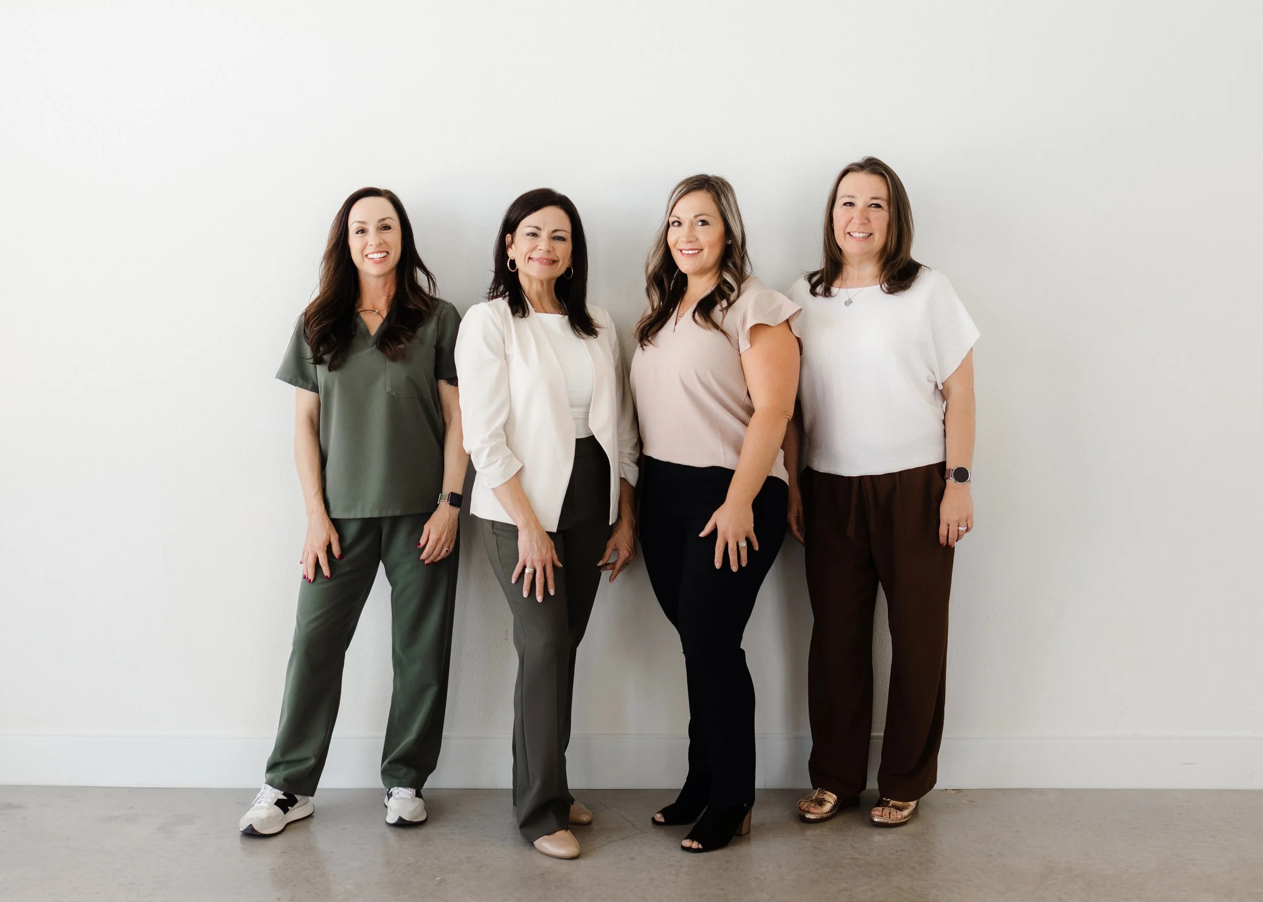 Four women standing side by side against a white wall, smiling and dressed in casual business/medical attire.