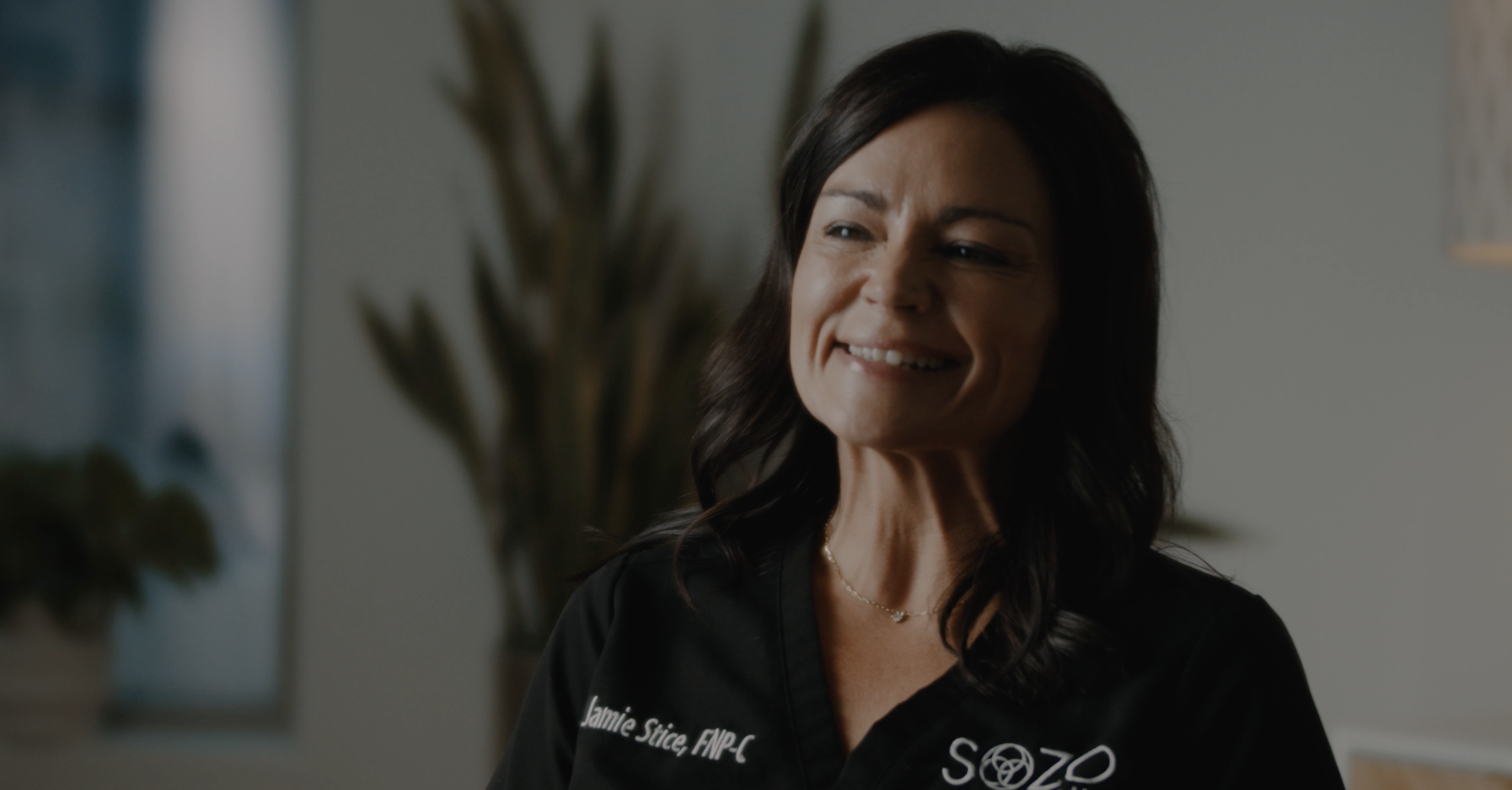 Jamie Stice, FNP-C | A woman with dark hair smiling, wearing a black scrub top with white text, in a room with a blurred plant in the background.