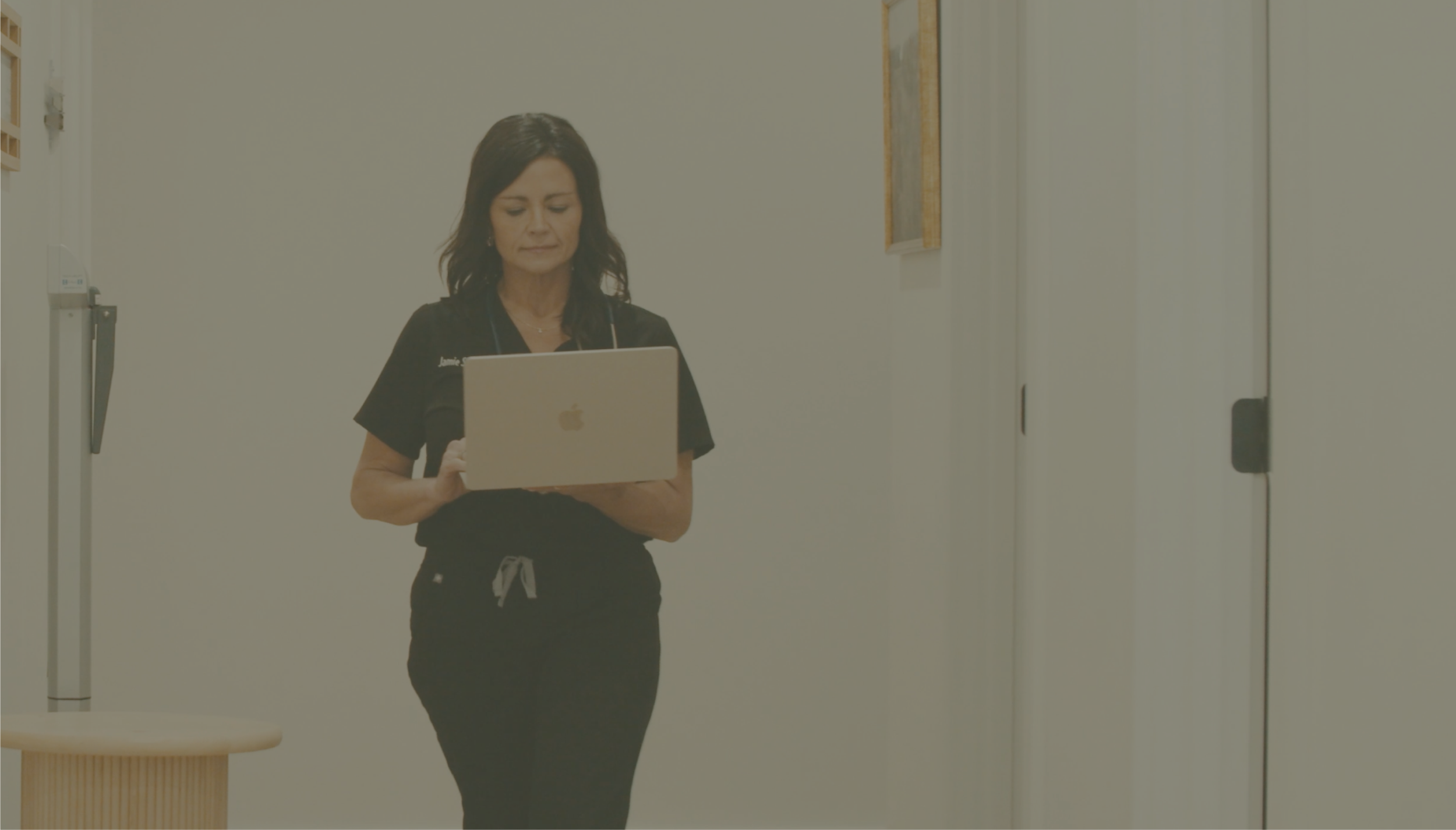 Jamie Stice, NPC | A woman with shoulder-length dark hair wearing black scrubs and stethoscope examines a laptop in a clinical setting.