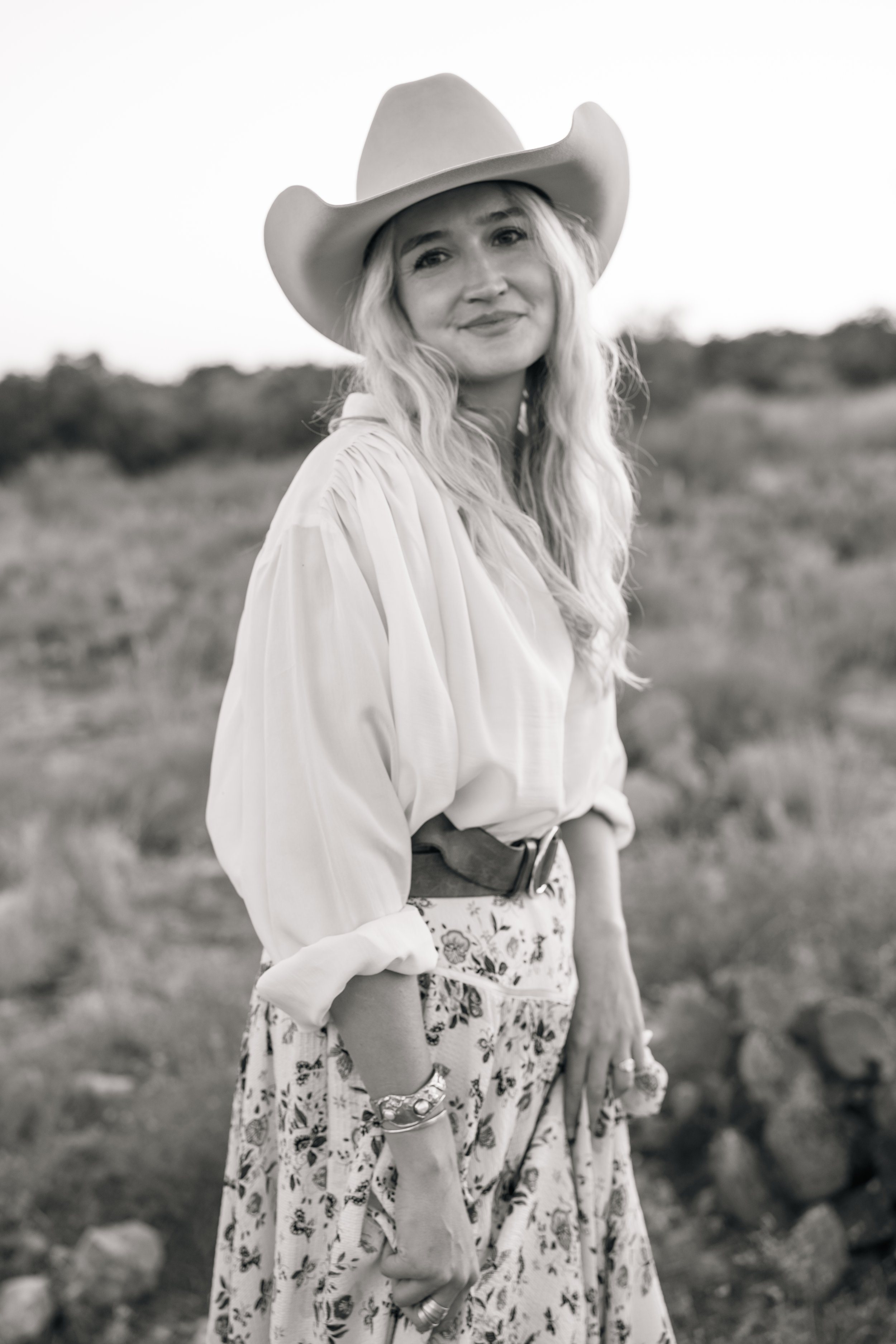 A woman with long wavy hair wearing a cowboy hat, a light-colored blouse, and a floral skirt, standing outdoors in a rural or countryside setting.