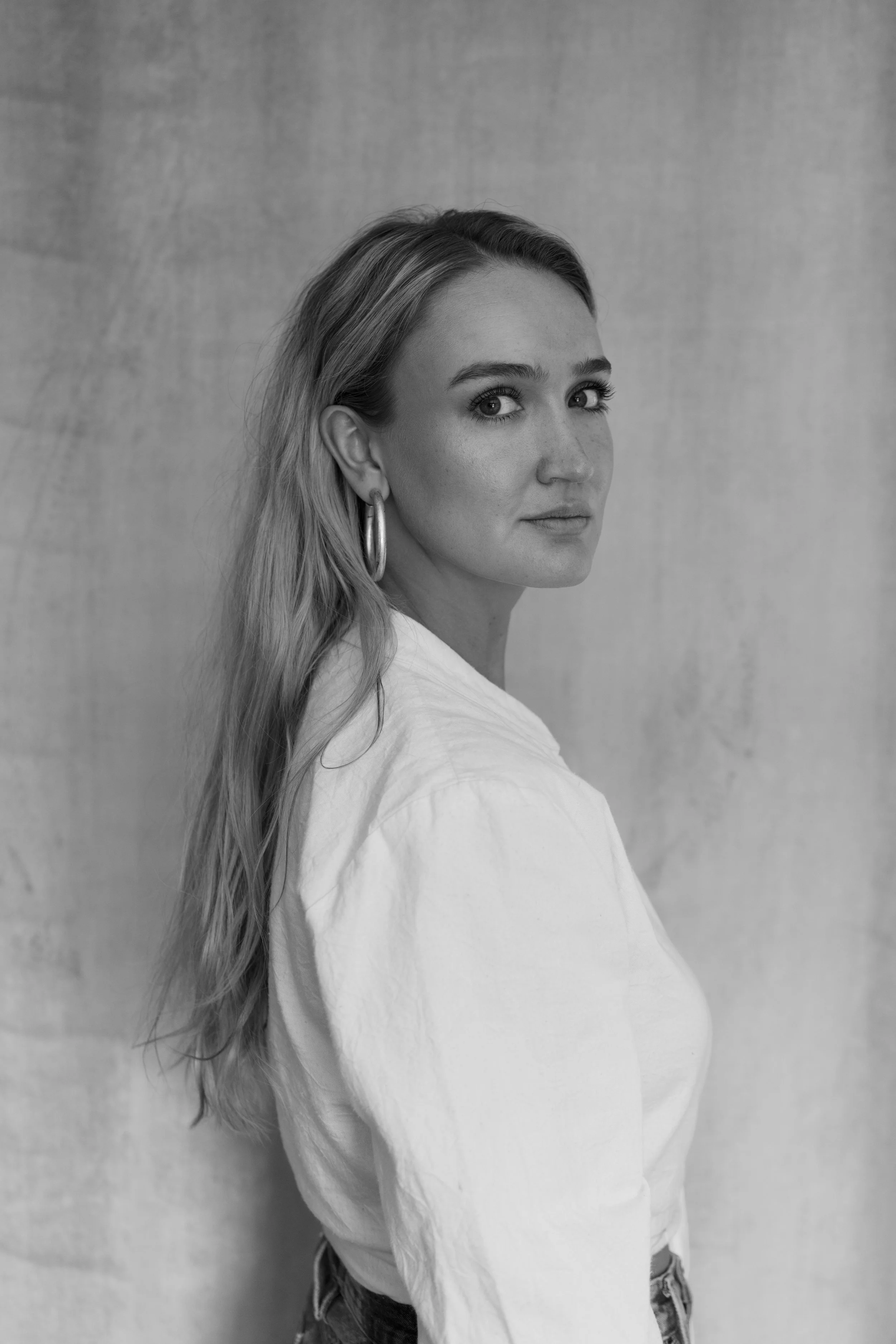 Black and white portrait of a woman with long wavy hair, wearing hoop earrings and a white top, standing against a plain background.