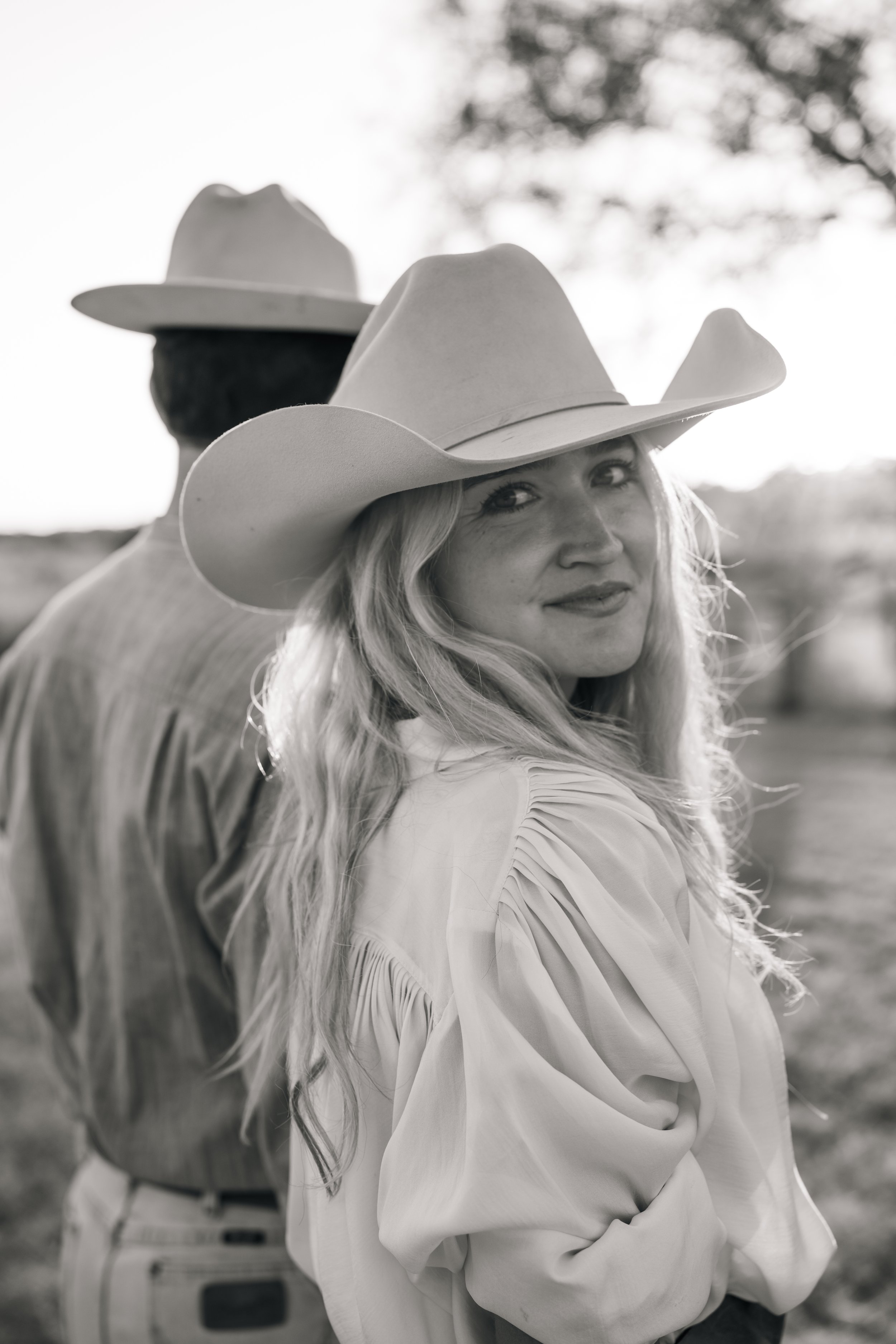 Black and white photo of a woman with long hair wearing a large cowboy hat, smiling, with a man in the background also wearing a cowboy hat, outdoors with trees in the background.
