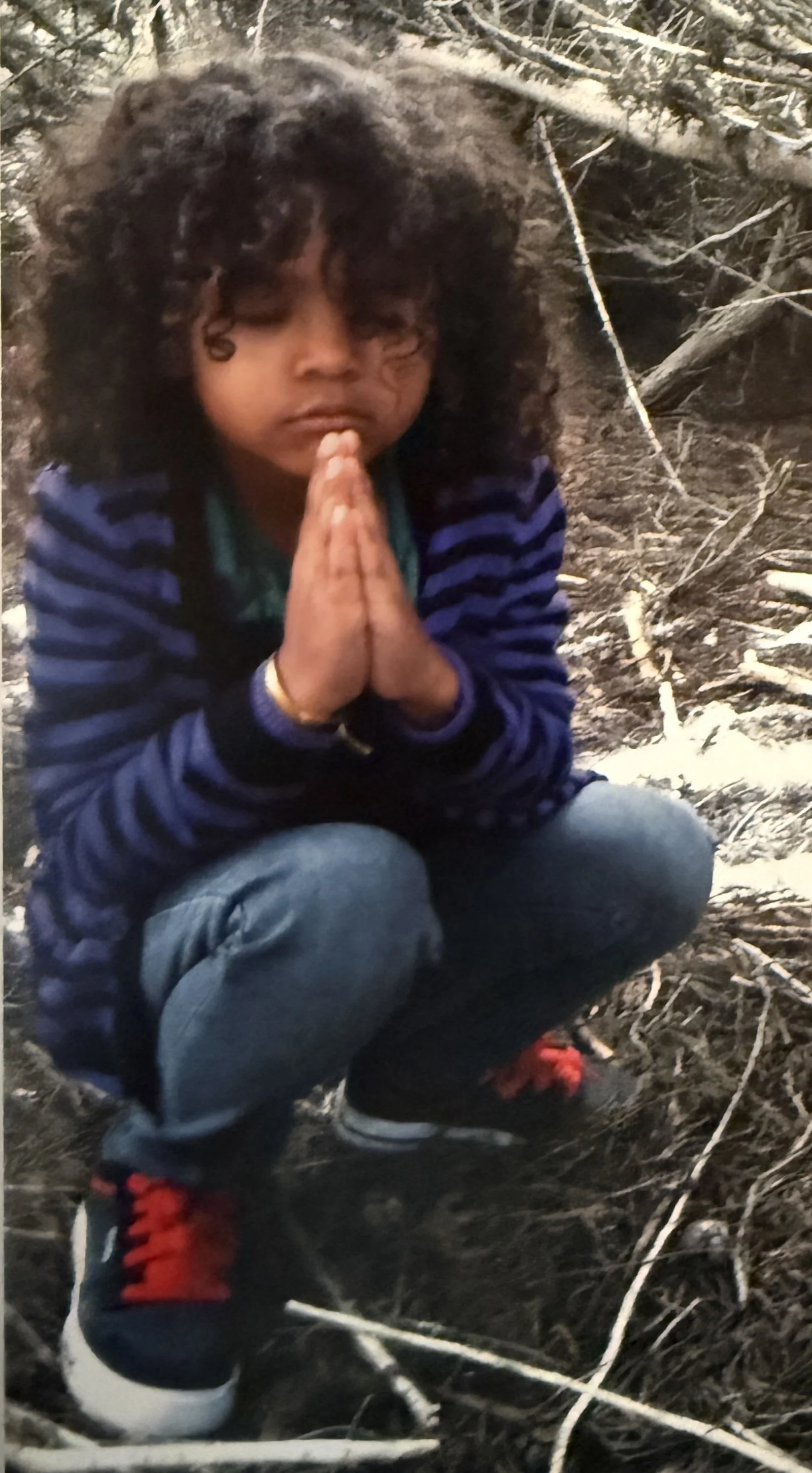 A young child with curly dark hair, dressed in a blue and black striped jacket, kneeling on muddy ground covered with twigs and branches, with hands pressed together in a prayer-like gesture.