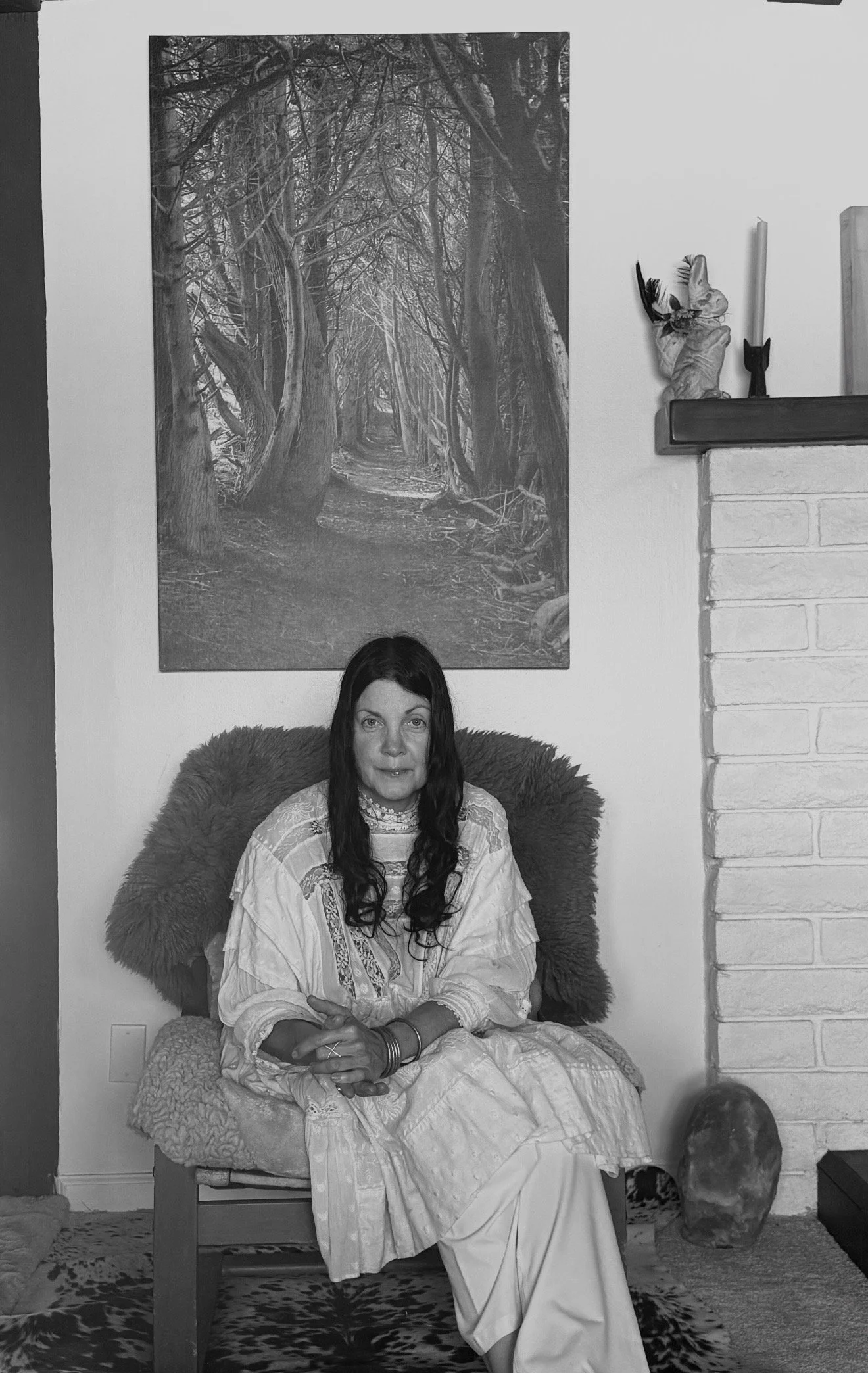 A woman with long dark hair sitting on a curly fur-covered chair in front of a white wall with a large black and white forest scene painting and decorative objects on a shelf on the wall. She is wearing traditional Indian clothing.
