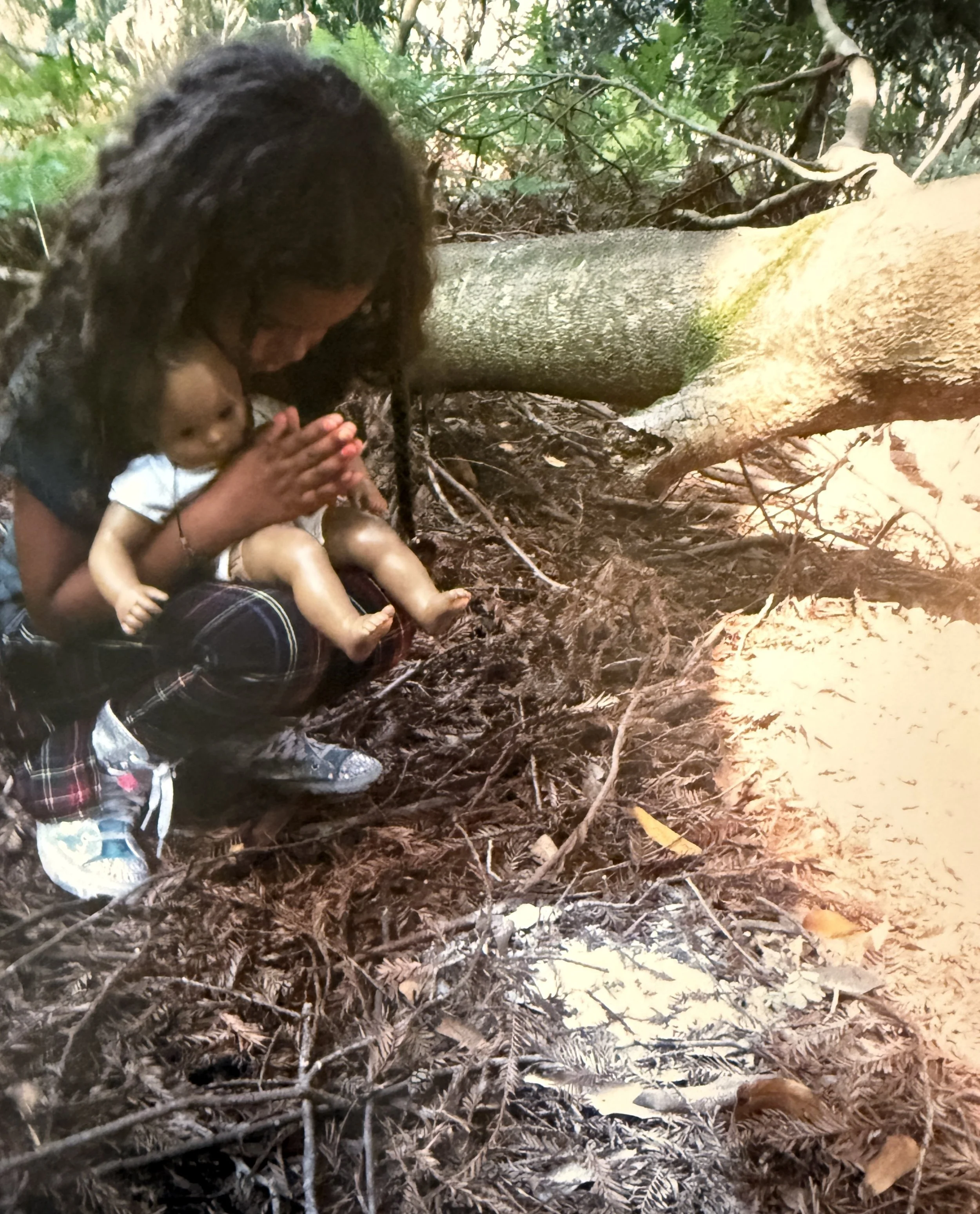 A woman sitting on the forest floor with a small child, surrounded by fallen leaves and branches, with a large fallen tree behind them.
