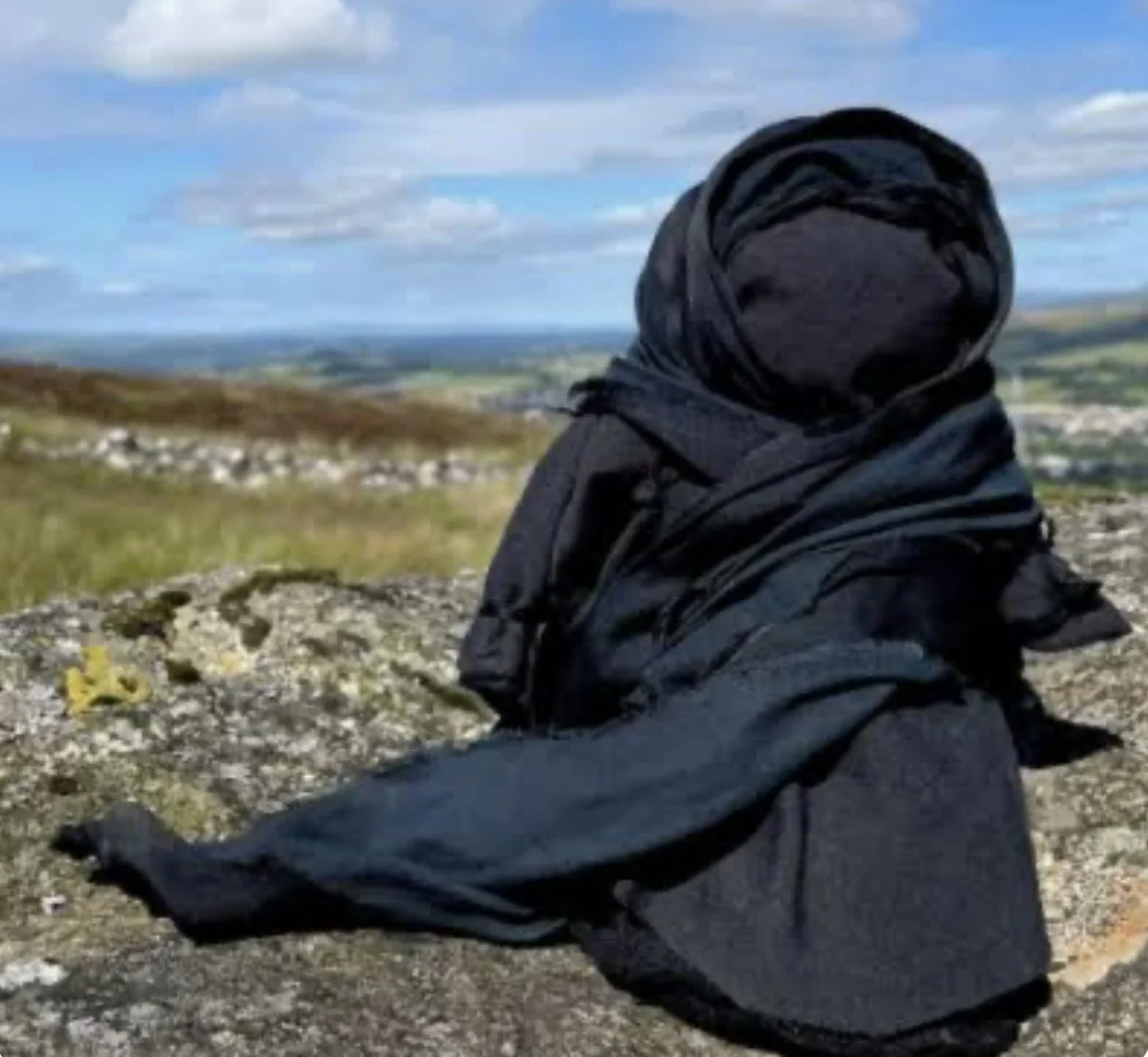 Person wearing a black headscarf and jacket sitting on a rocky hilltop, overlooking a wide landscape with fields, a stone wall, and a partly cloudy sky.